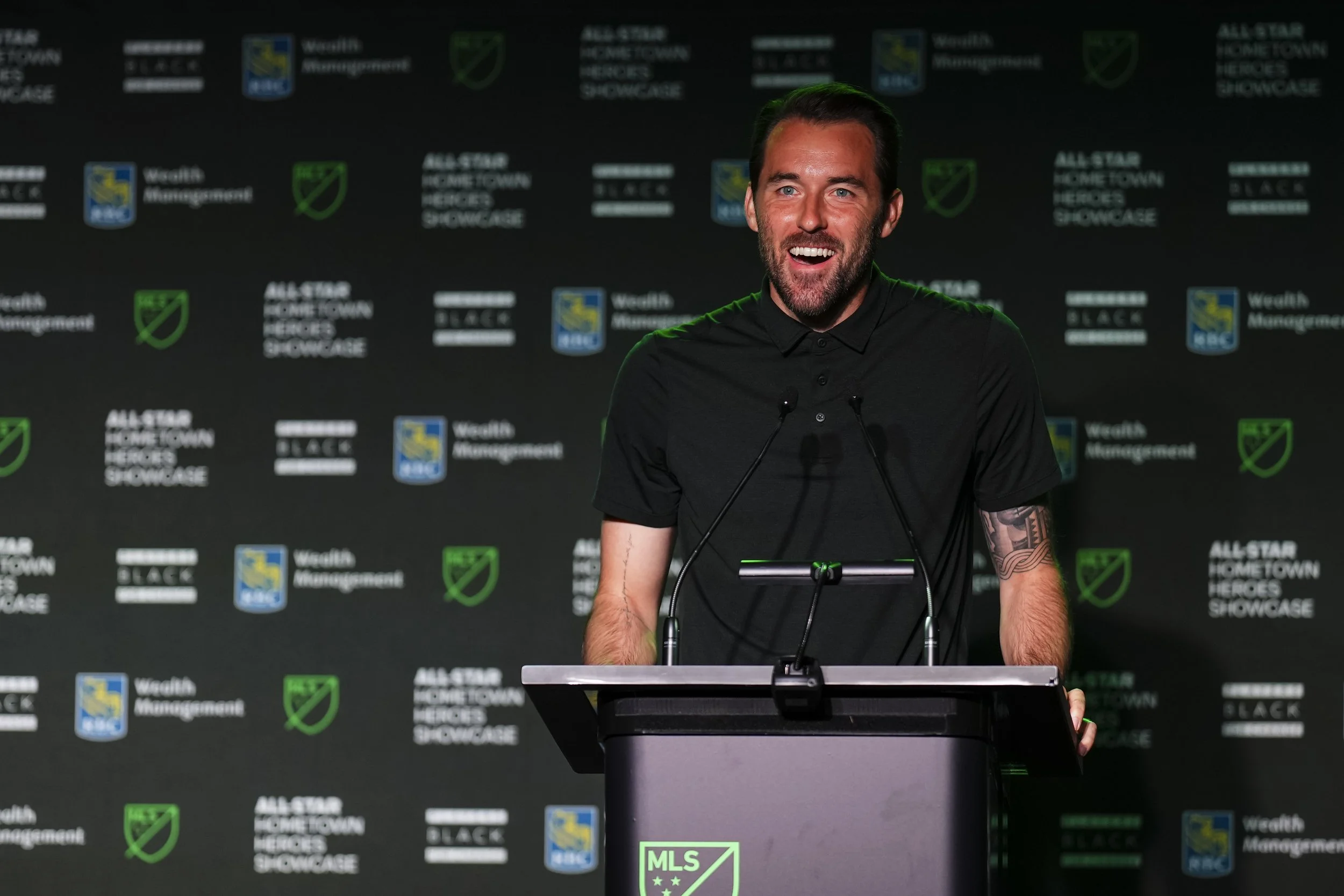A man at a podium giving a speech at the MLS All-Star Home Town Heroes Showcase event. He has a beard, is smiling, and is wearing a black polo shirt. There are microphones in front of him and a backdrop with MLS and W Columbus Management logos.
