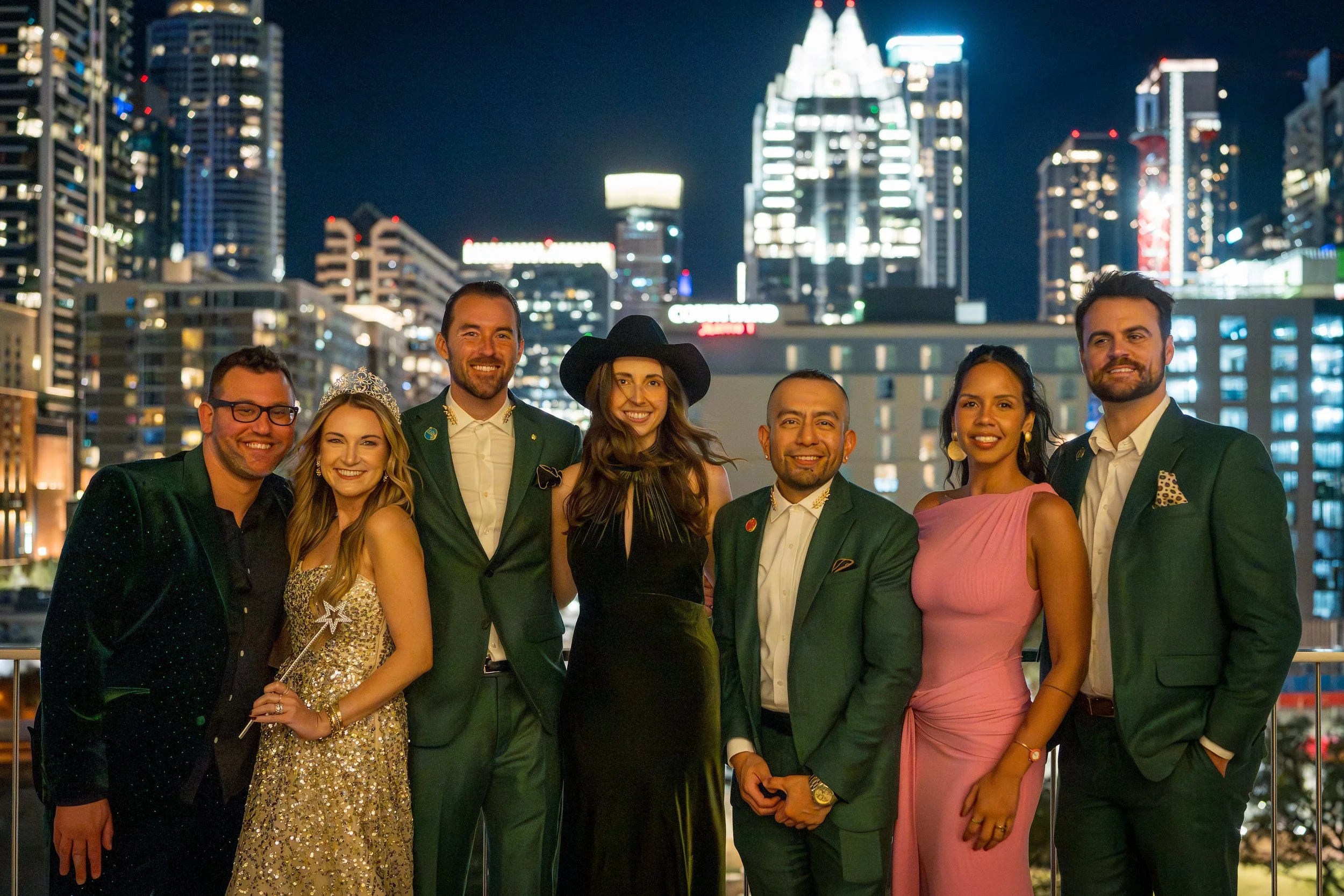 Group of eight diverse people dressed in fancy clothes, standing on a balcony with a city skyline at night in the background.