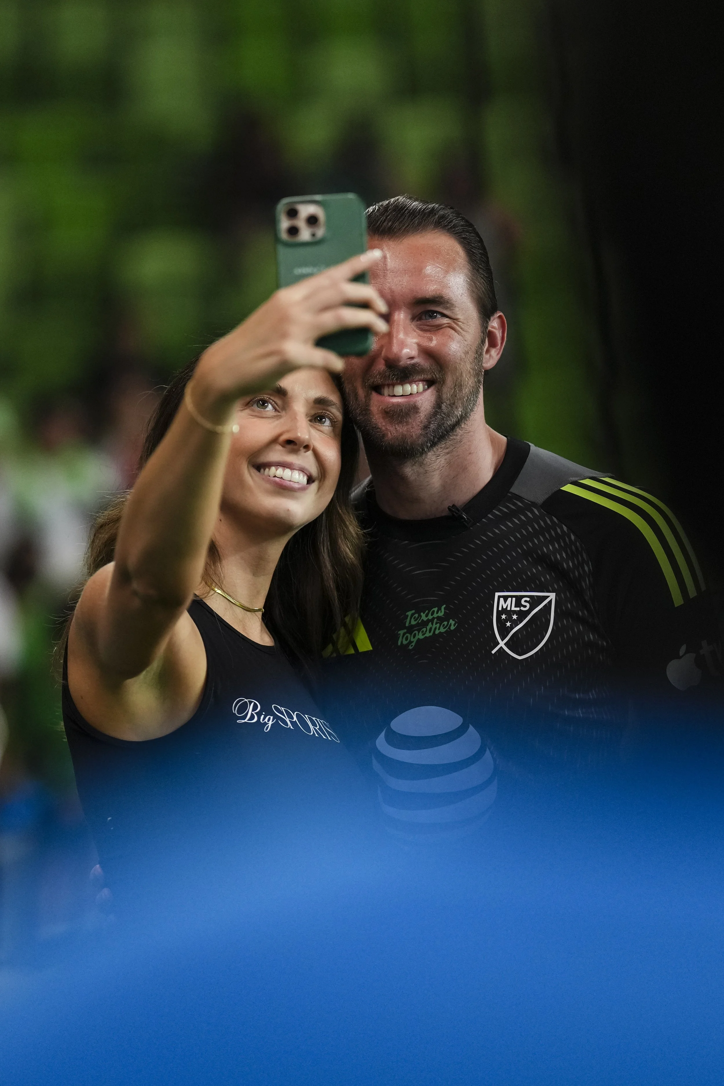 A woman and a man taking a selfie together, smiling. The woman has long brown hair and is wearing a black Top. The man has short dark hair and a beard, wearing a black MLS soccer jersey with green accents. They are in a green wooded outdoor setting.