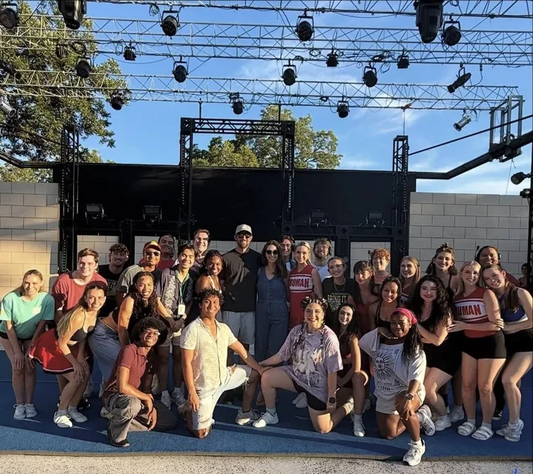 A group of people, including children and adults, posing together outdoors in front of a stage with rigging and lights, on a blue track or sports field, during daytime.