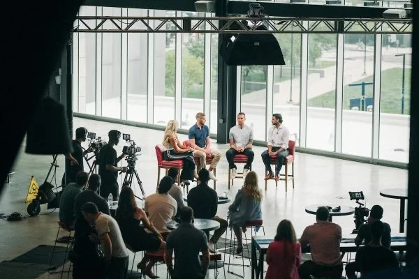 Panel discussion with four speakers seated on chairs in front of a large glass wall, audience watching, recording equipment visible, in a modern studio setting.