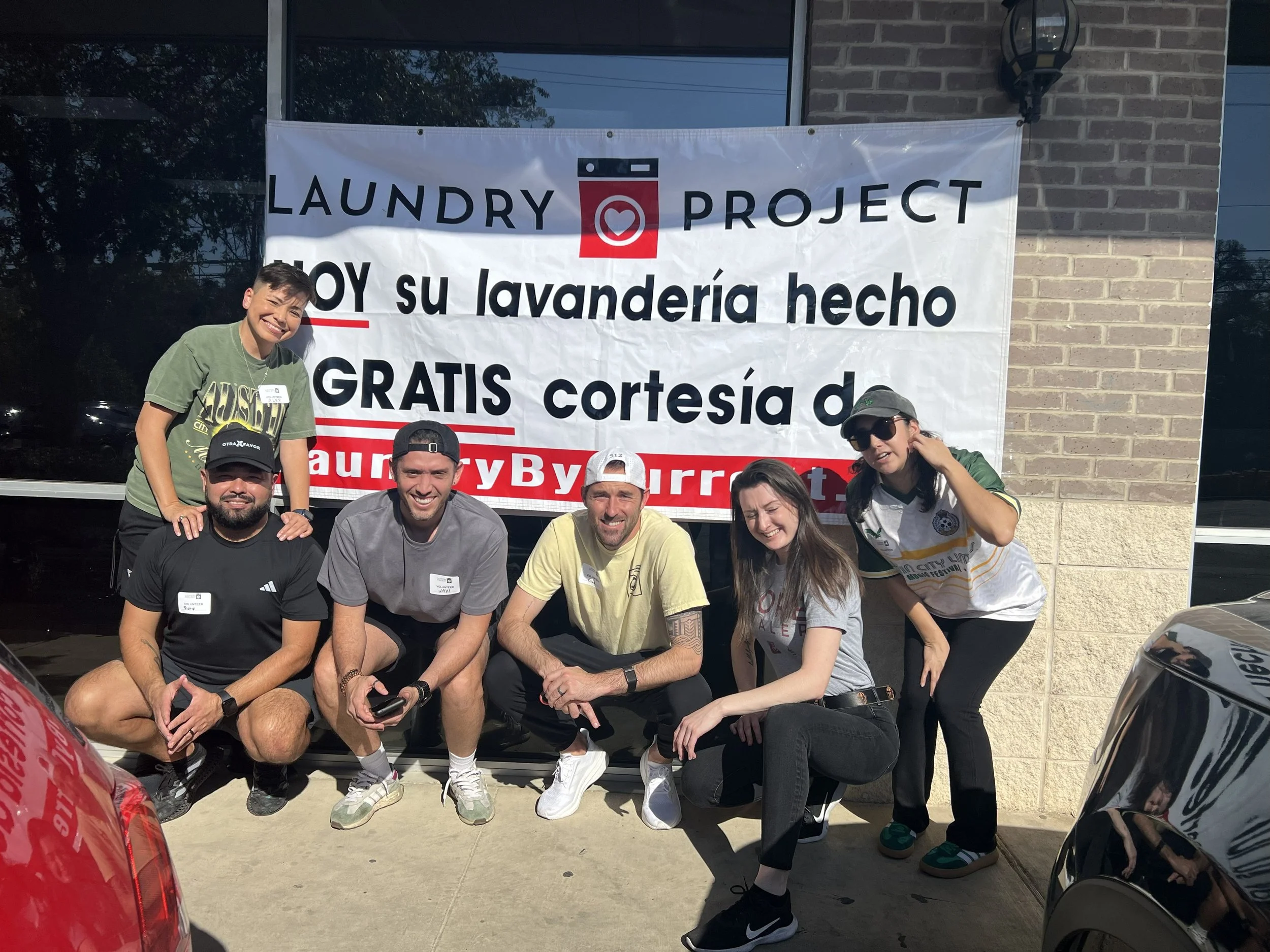 Group of six people smiling and posing in front of a banner related to a laundry project, outside of a building with a brick wall and windows.
