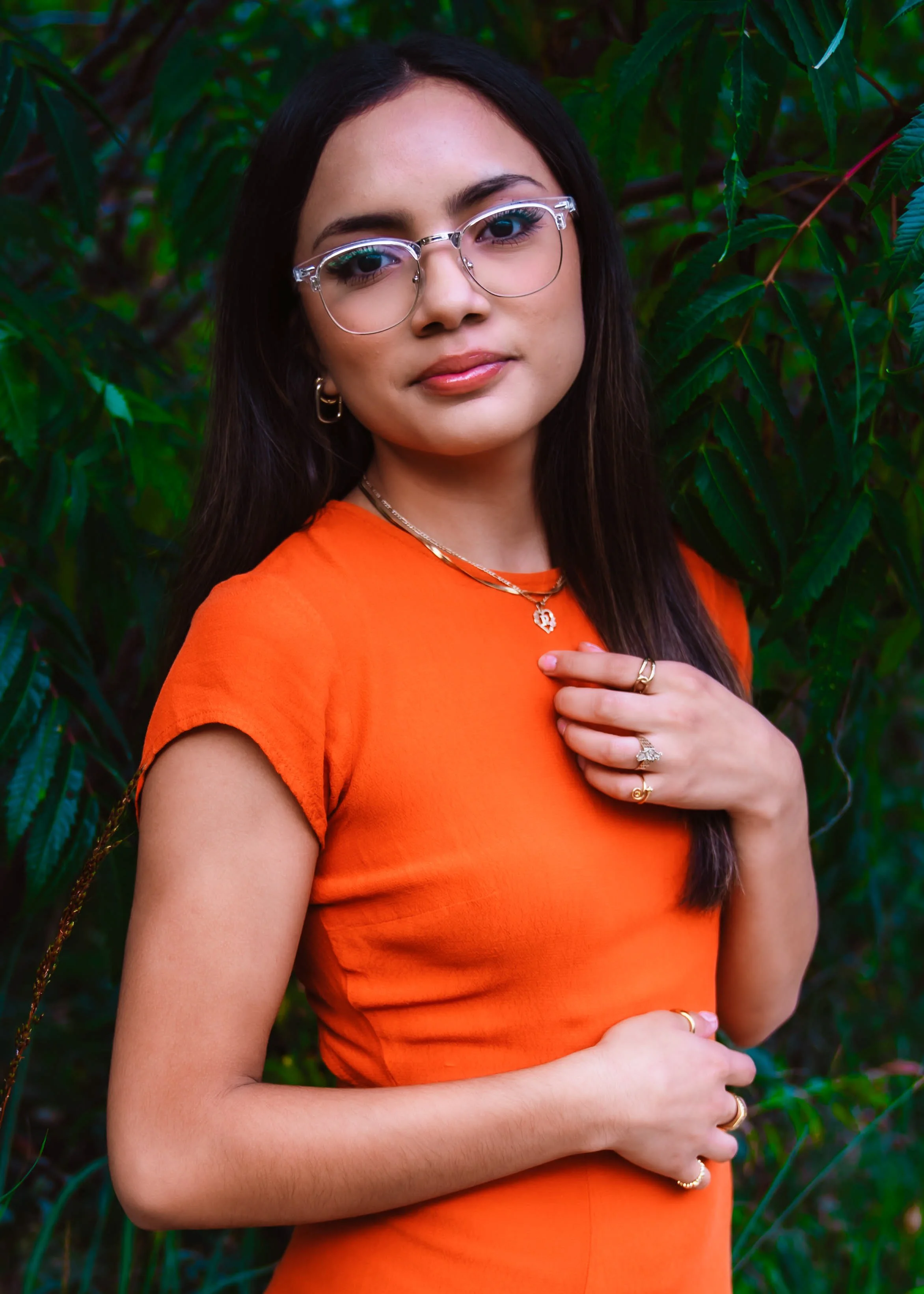 A young woman with long dark hair, glasses, and earrings, wearing an orange top and gold jewelry, standing outdoors in front of green foliage.