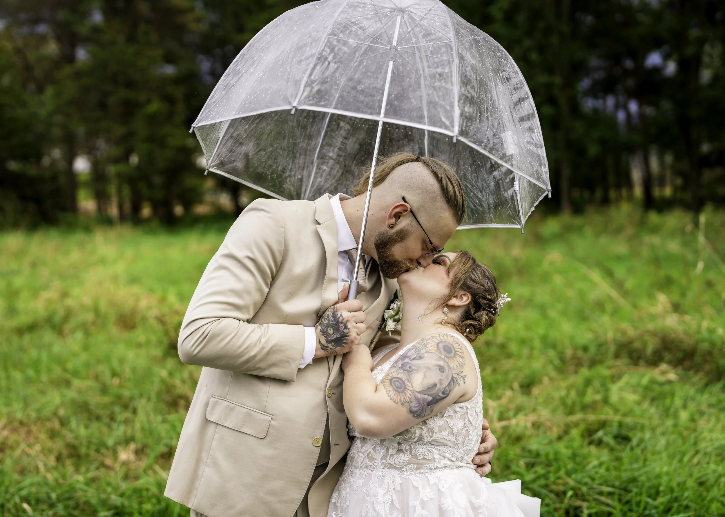 A couple kissing under a transparent umbrella outdoors on a rainy day, with green grass and trees in the background.