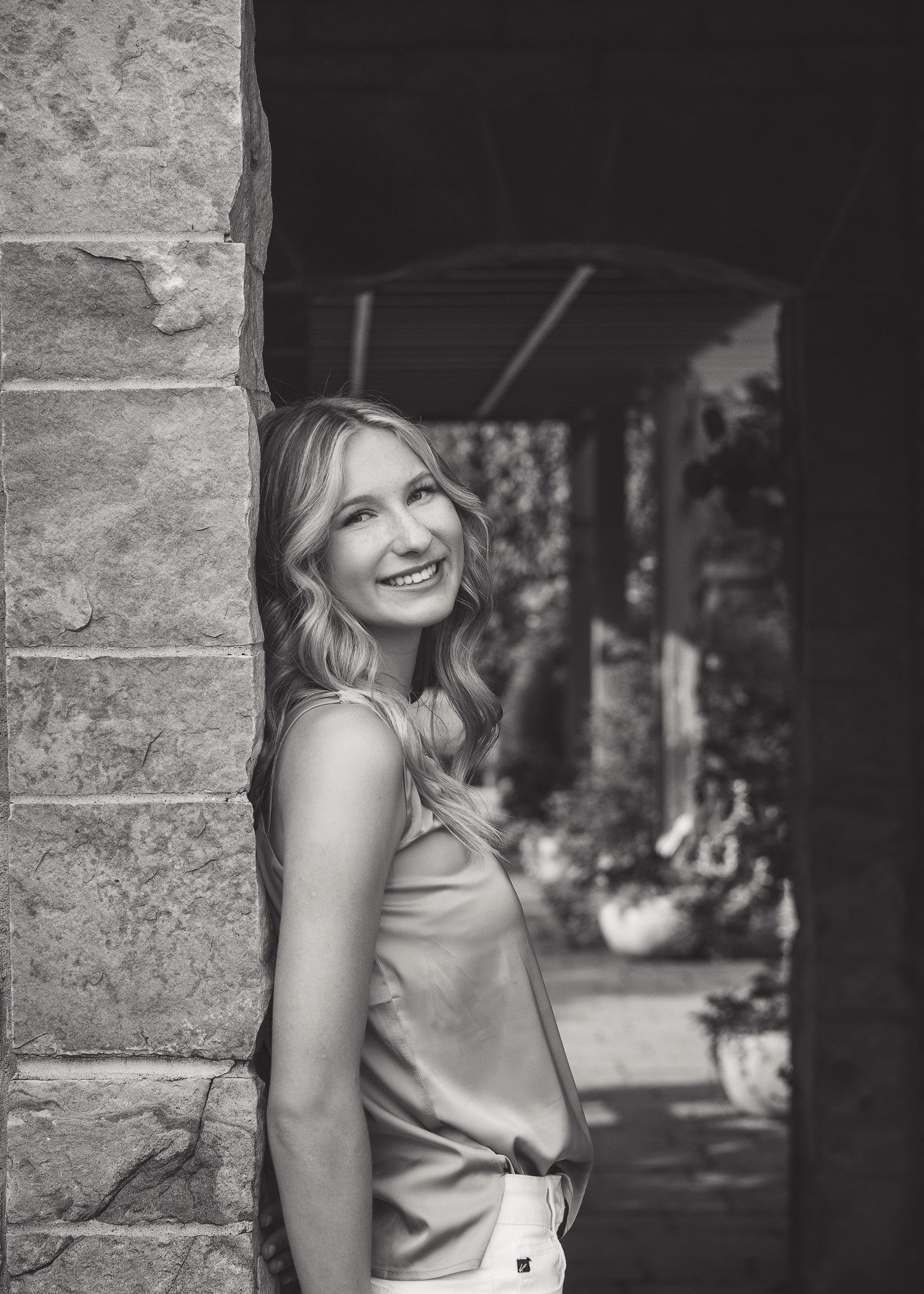A young woman with wavy hair smiling, leaning against a stone wall in an outdoor setting, in black and white.