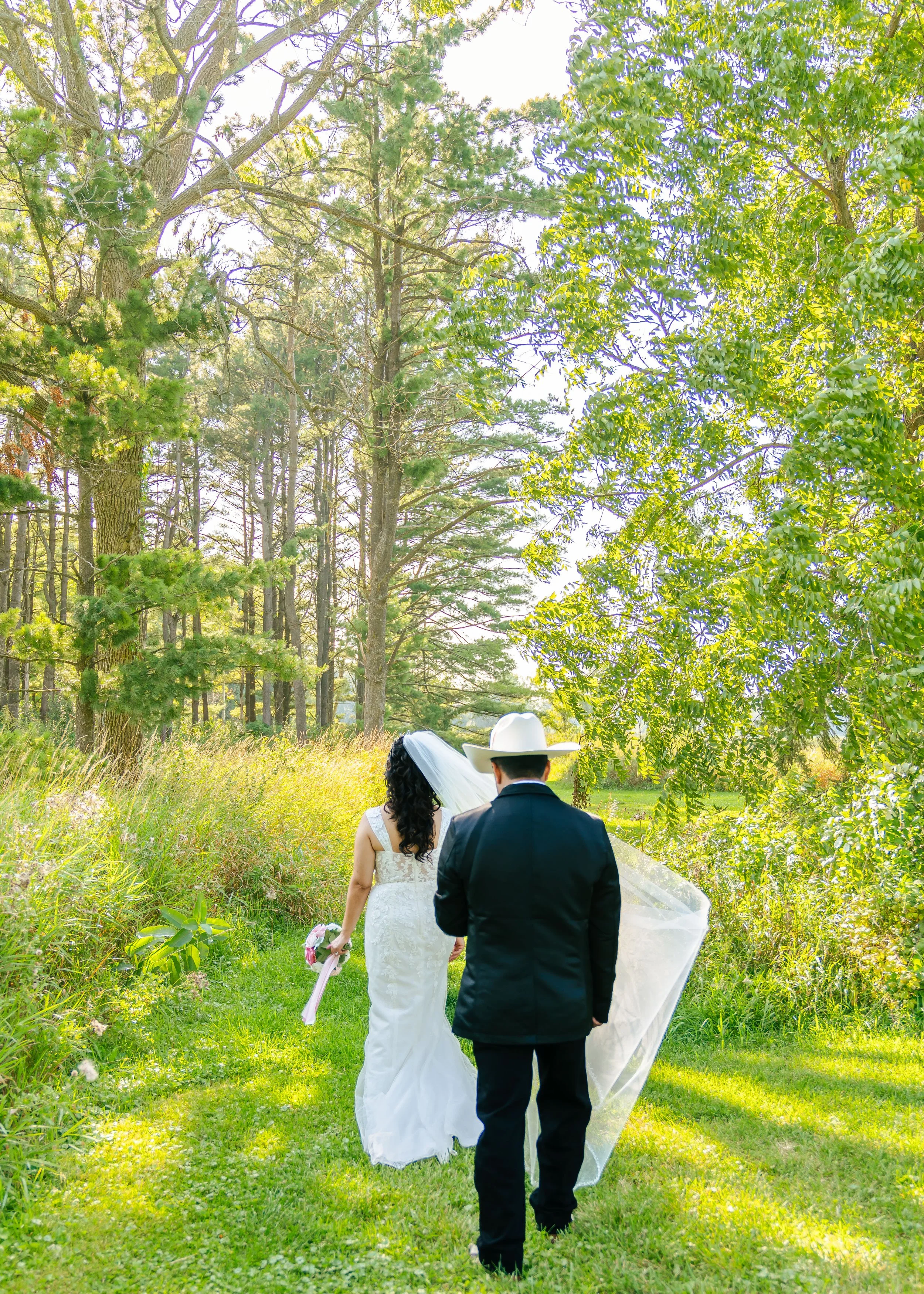 A bride and groom walking on a grassy path through a lush, green wooded area during a wedding photoshoot.