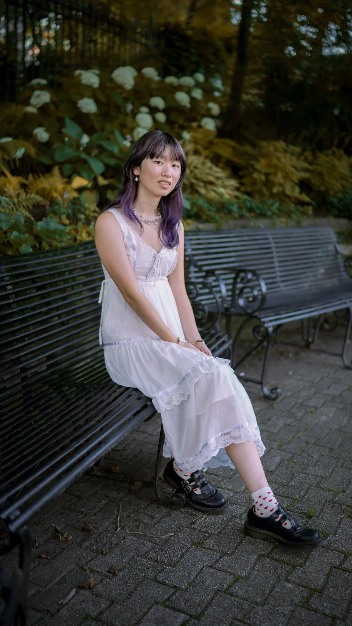 A young woman in a white dress with purple hair sitting on a black metal park bench in a garden with white flowers and green foliage in the background.