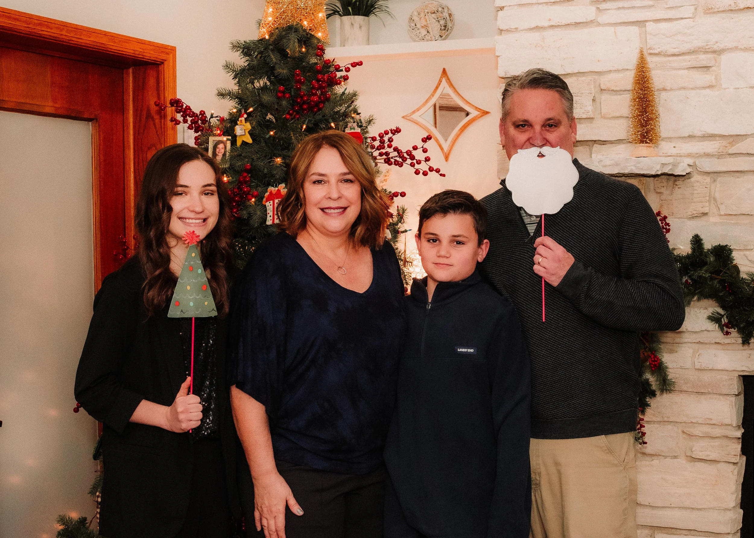 A family of five celebrating Christmas indoors, standing in front of a decorated Christmas tree with red ornaments and lights, and a white brick fireplace with holiday decorations.