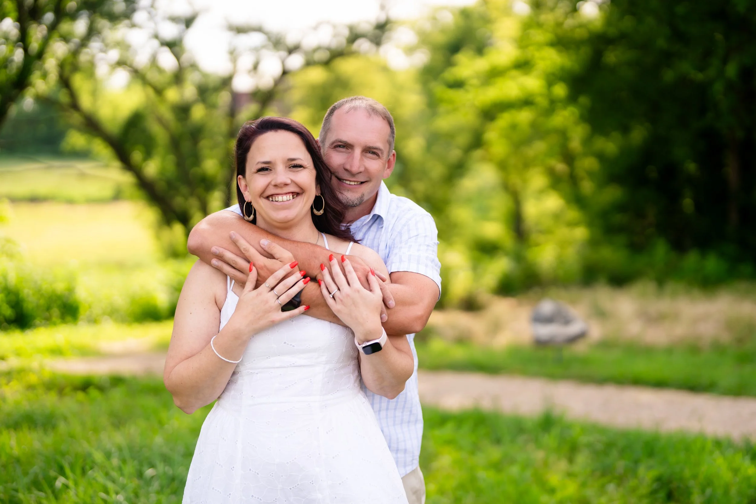 A smiling couple embracing outdoors, the woman wearing a white dress and the man in a light-colored shirt, surrounded by green trees and grass.