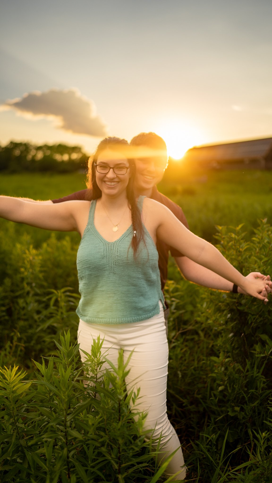 Two people, a woman in a sleeveless green top and white pants and a man, are standing in a field of green plants during sunset, smiling and holding hands. The sun is setting behind them, casting a warm glow.
