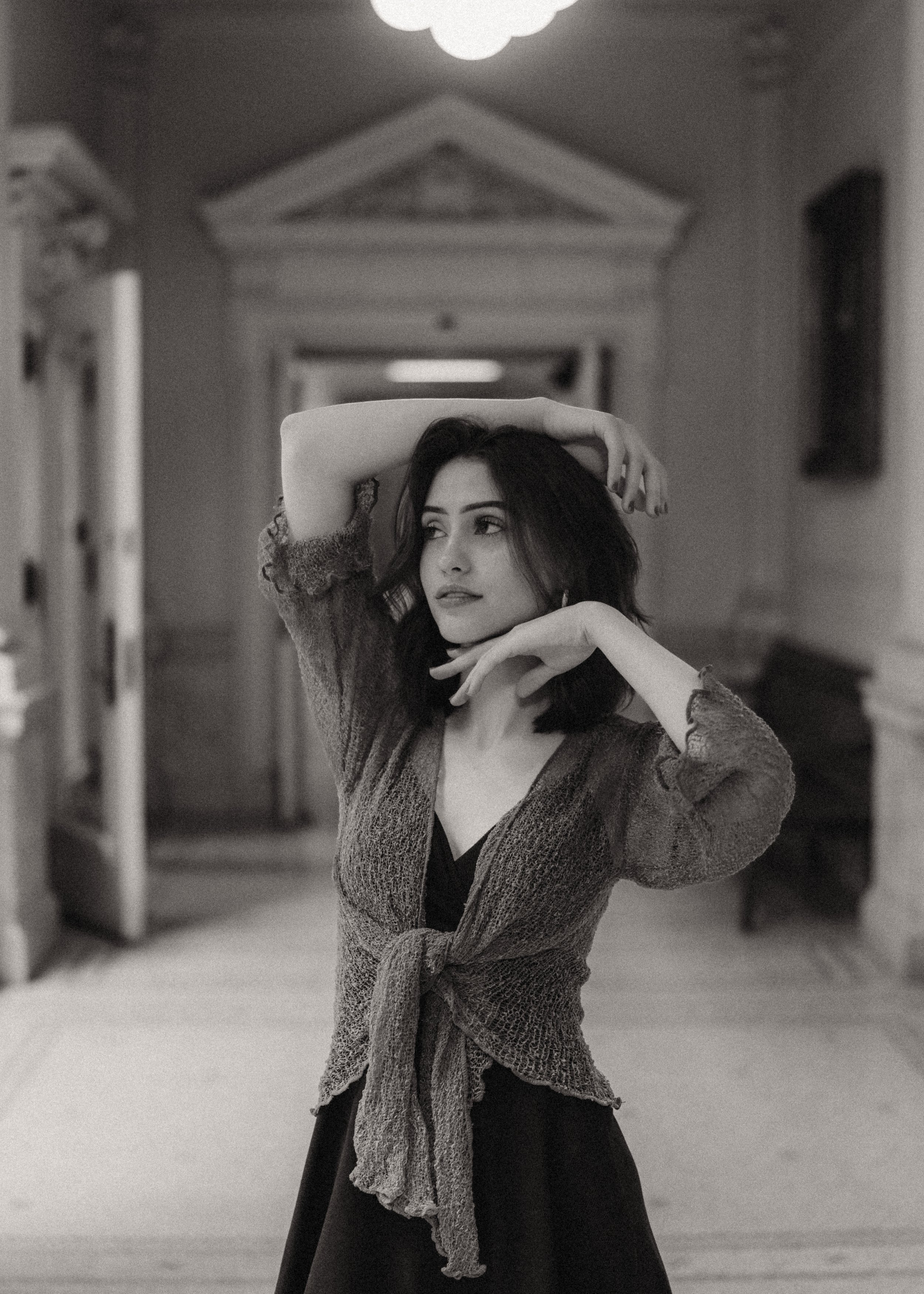 A woman with dark hair and expressive eyes poses with her hands above her head and near her face in an elegant hallway.
