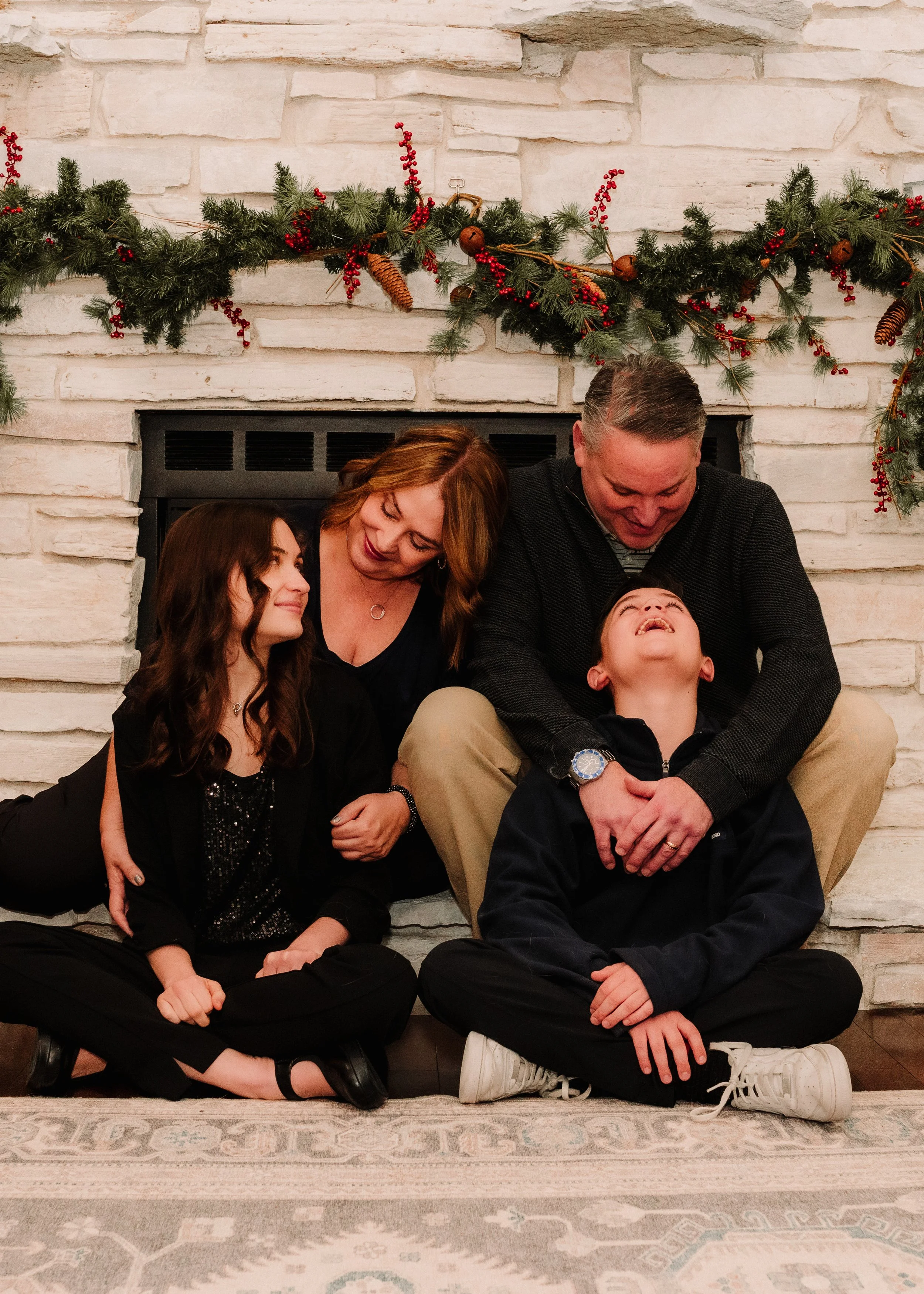 A family of four sitting on a rug in front of a fireplace decorated with a Christmas garland. The father and son are laughing, while the mother and daughter are smiling at each other.