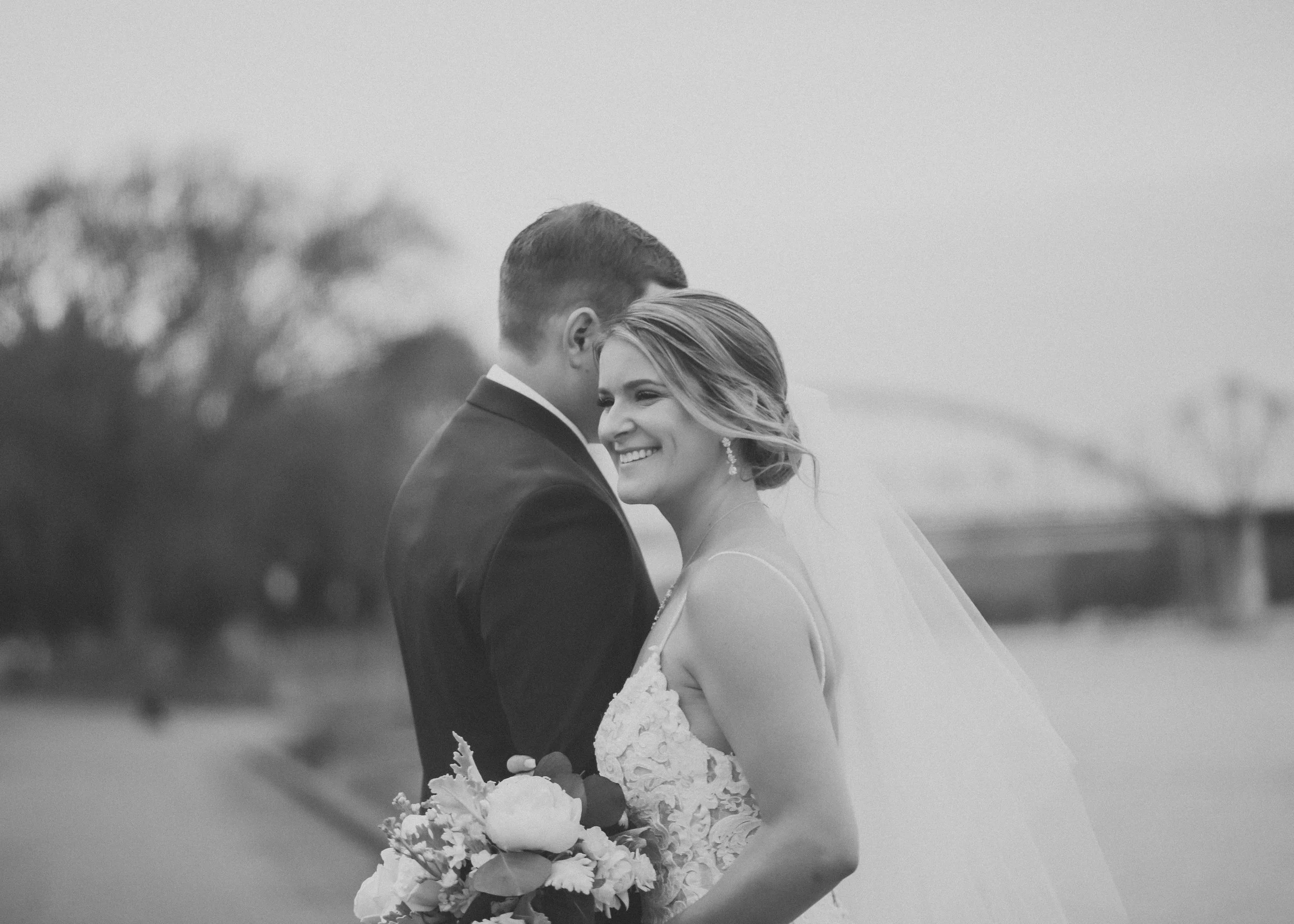 Black and white photo of a bride and groom embracing outdoors, with the groom's face close to the bride's head, both smiling, near a river or body of water.
