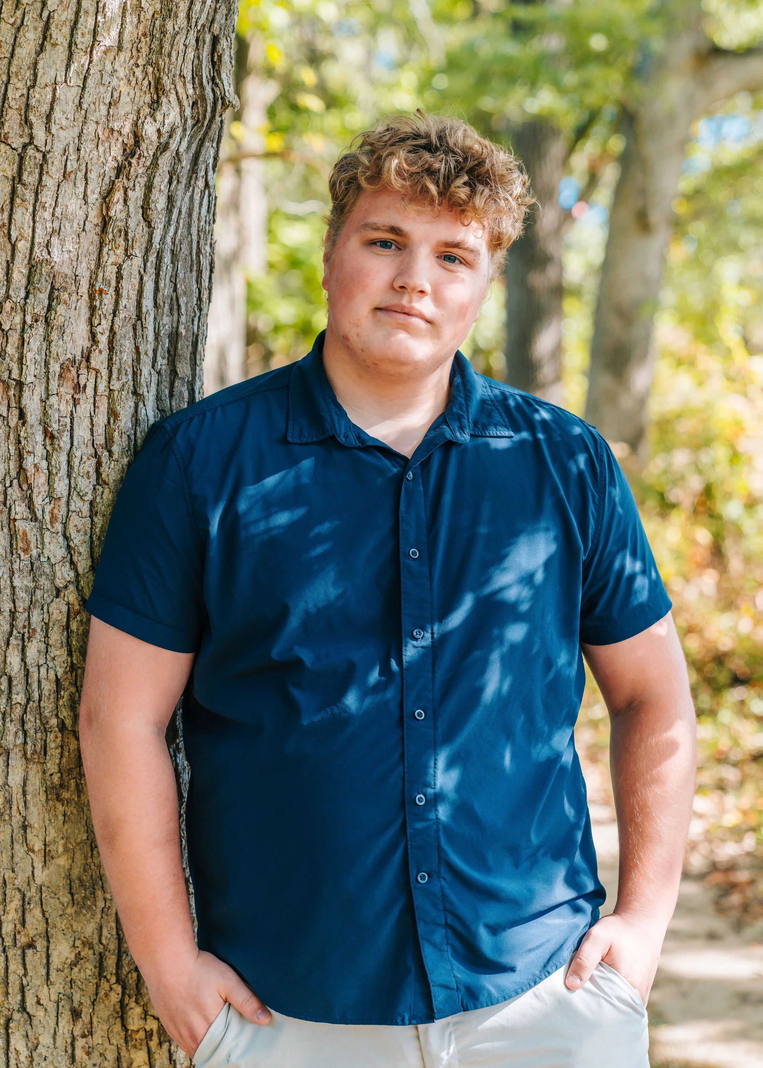A young man with curly hair leaning against a tree in a forest, wearing a navy blue short-sleeved button-up shirt and light-colored pants.