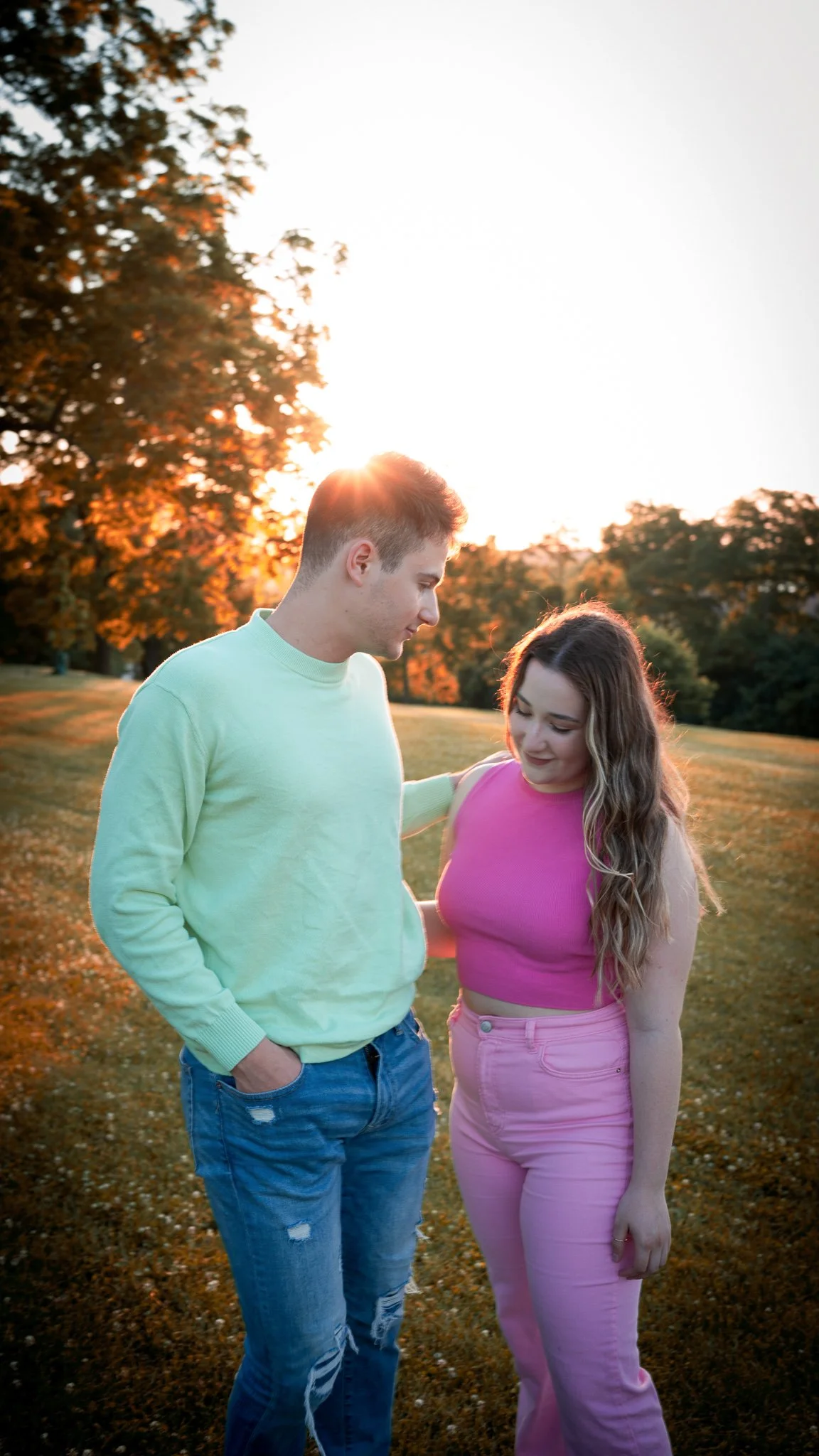 A young man and woman standing in a park during sunset, looking at each other. The man is wearing a light green sweater and ripped jeans, the woman is wearing a pink sleeveless top and pink pants, with trees in the background.