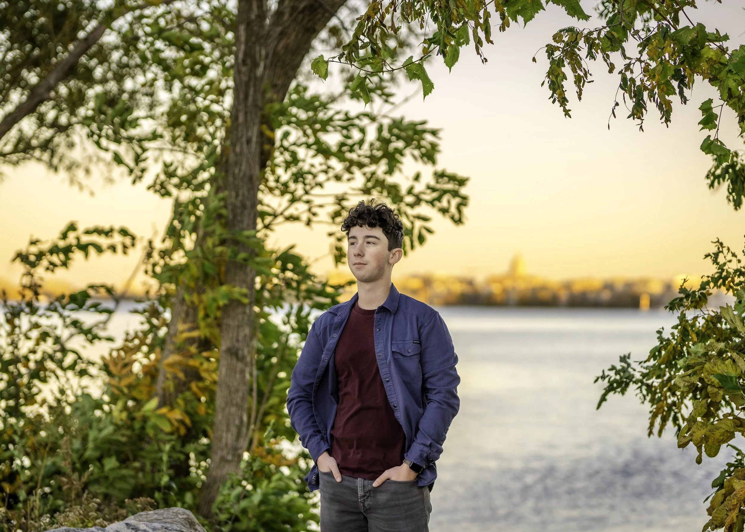 A young man with dark curly hair wearing a blue jacket and maroon shirt standing outdoors near a body of water during sunset, with trees and a city skyline in the background