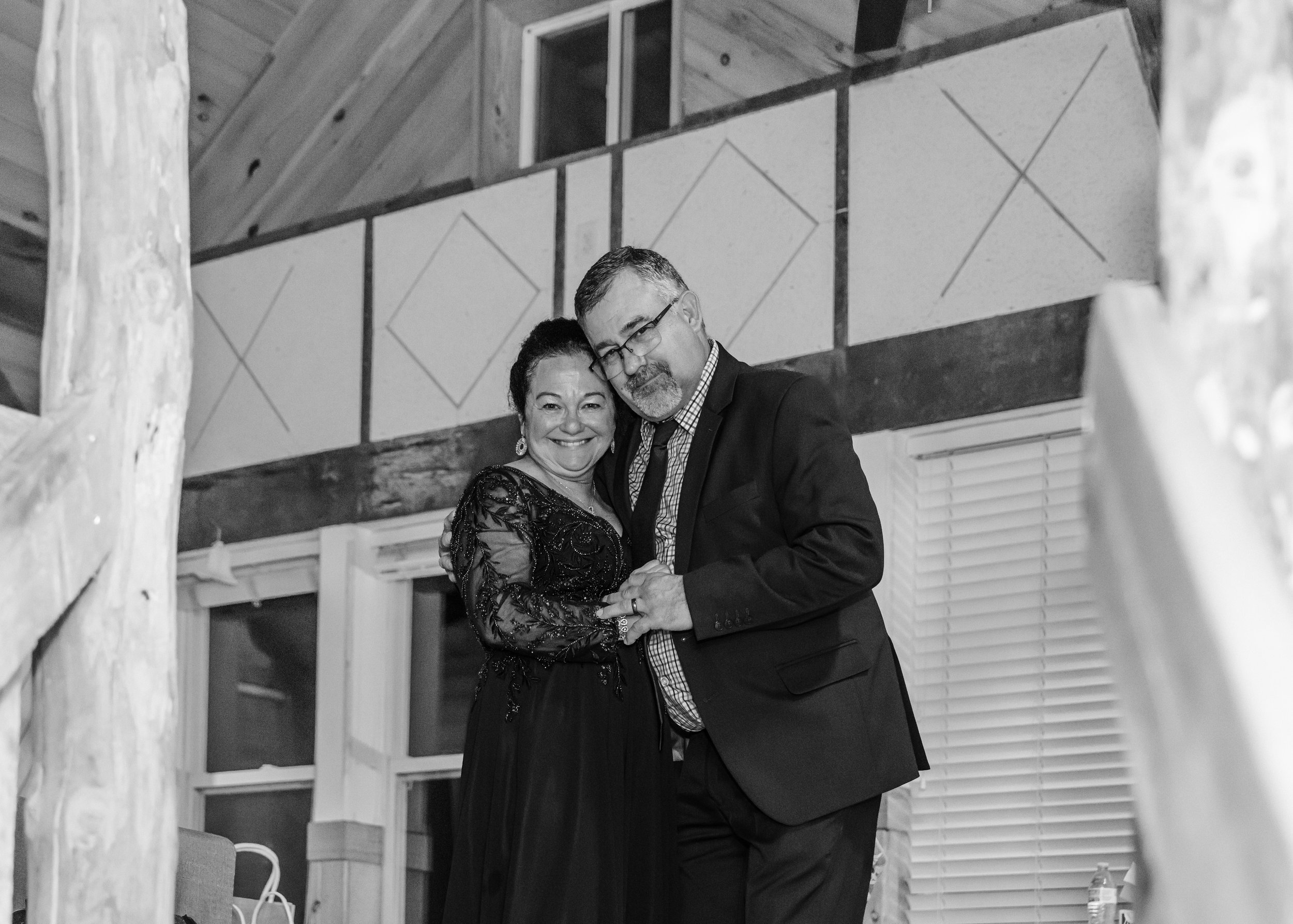 A smiling woman and a man in glasses, dressed in formal attire, dancing together indoors with rustic wooden beams and decorative wall panels in the background.