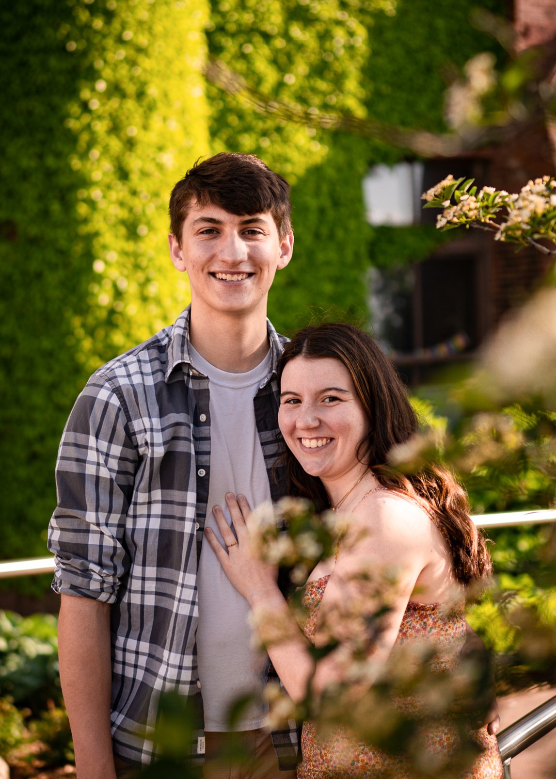 A smiling young man with dark hair in a plaid shirt standing next to a smiling young woman with long dark hair in a floral dress. They are outdoors with a lush green hedge and flowering trees in the background, enjoying warm sunlight.