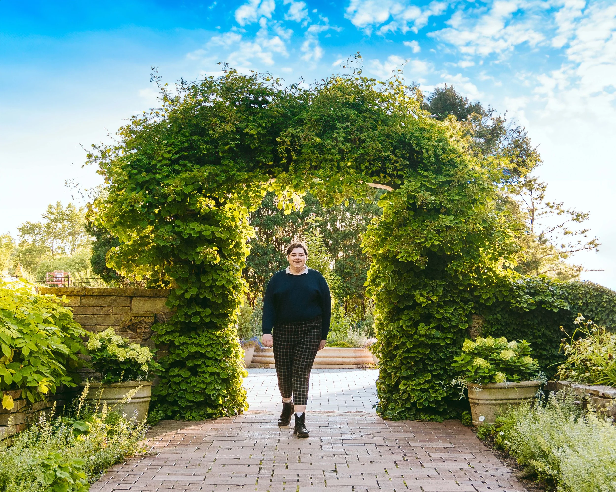 A person walking through a garden archway formed by lush green plants on a sunny day with a blue sky and scattered clouds.