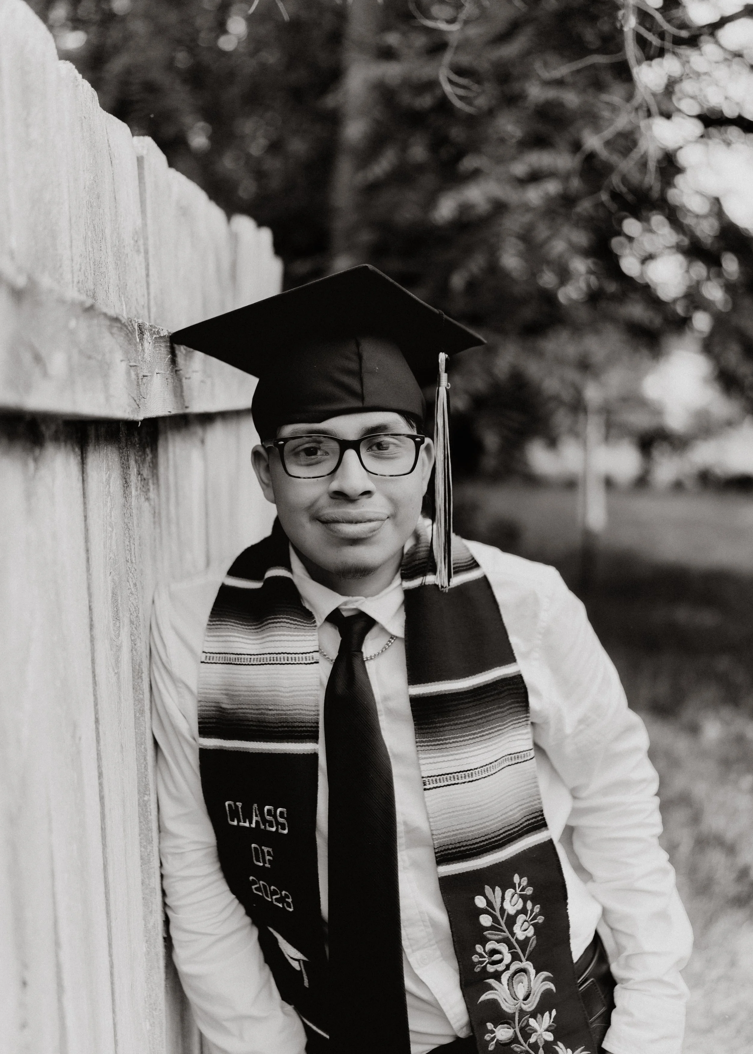 A young man wearing glasses, a graduation cap, and a stole with floral embroidery and 'Class of 2023' text, leaning against a wooden fence outdoors.