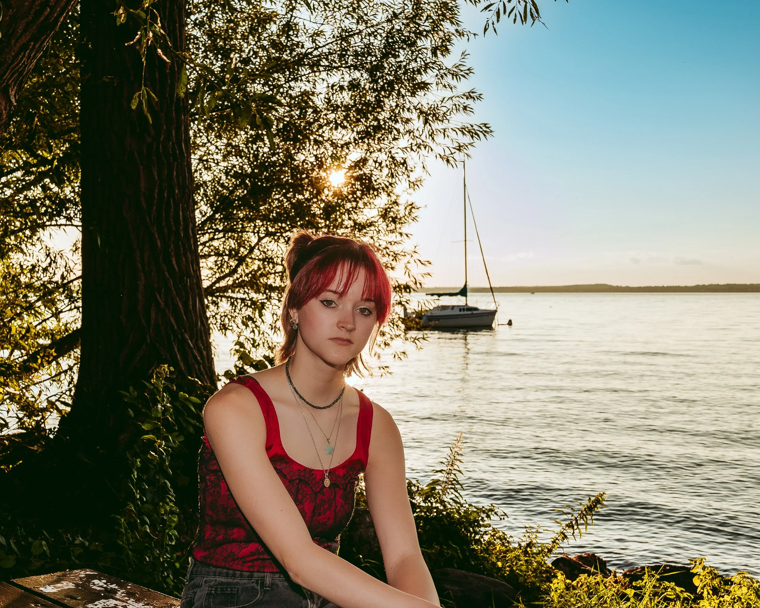 A young woman with red hair and dark eye makeup sitting outdoors near a body of water at sunset, with a sailboat in the background, surrounded by trees and bushes.