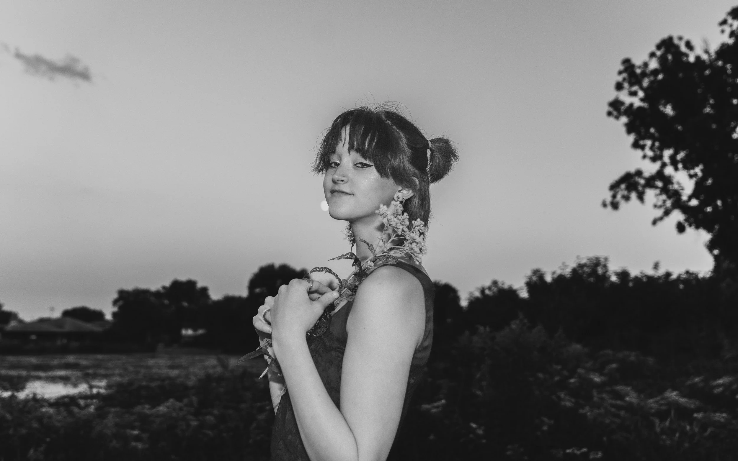 A woman with short hair styled in pigtails holding a flower, standing outdoors during dusk with trees in the background, in black and white.
