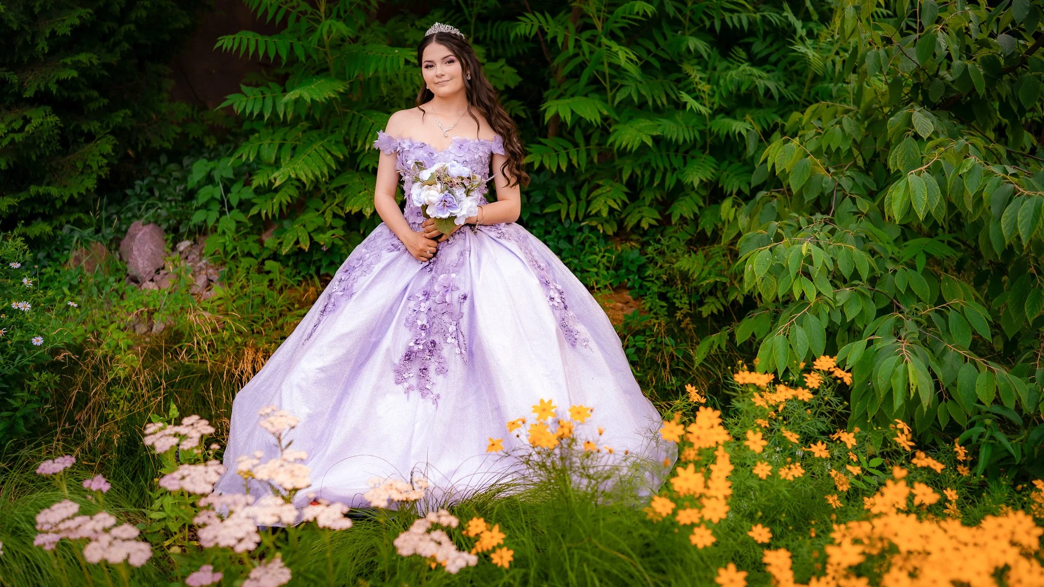 Young woman in a lavender quinceañera gown with floral embellishments, holding a bouquet, standing among colorful flowers and green foliage in an outdoor garden.