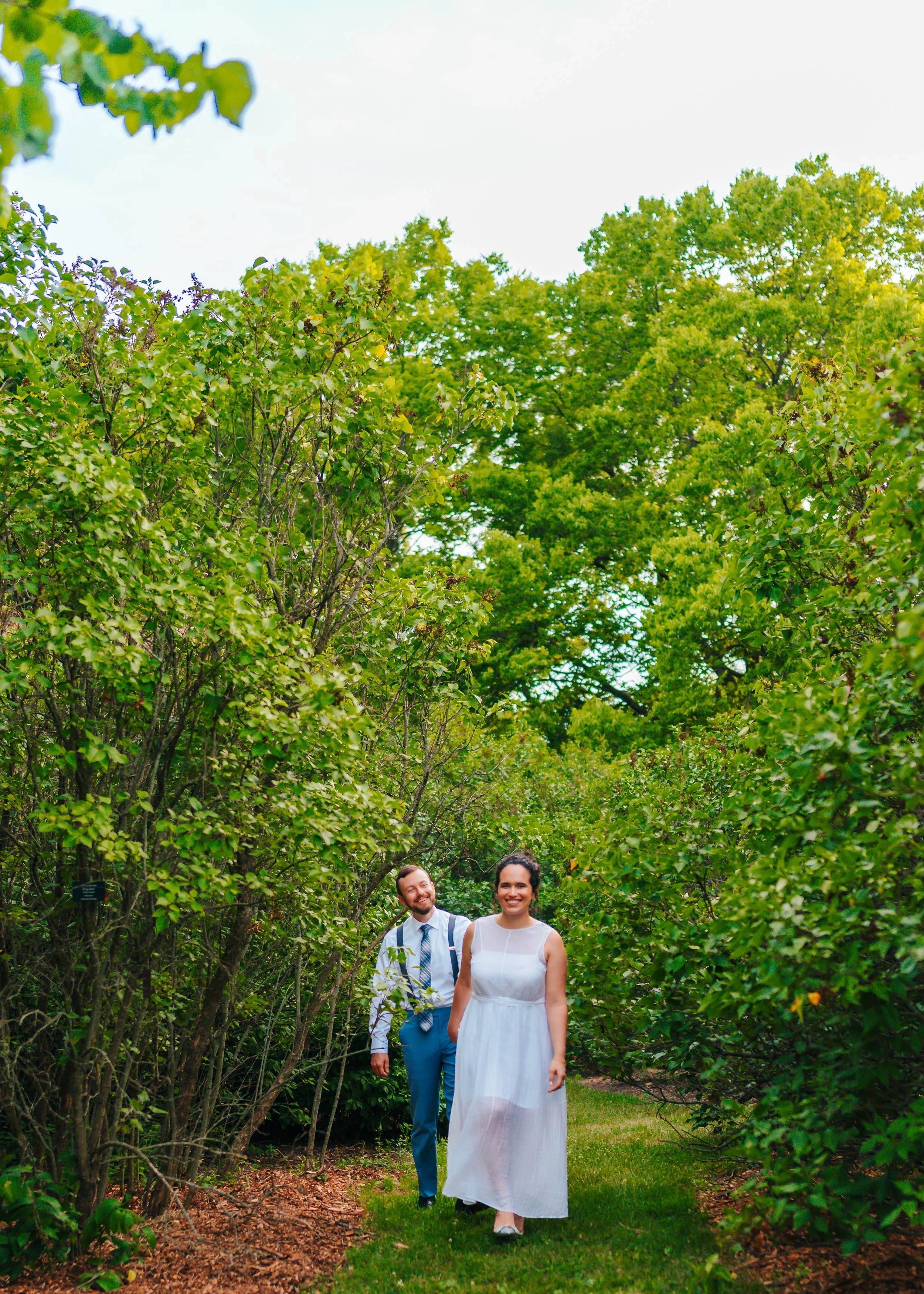 A man and woman walking through a lush green garden pathway, smiling, with trees and bushes surrounding them.