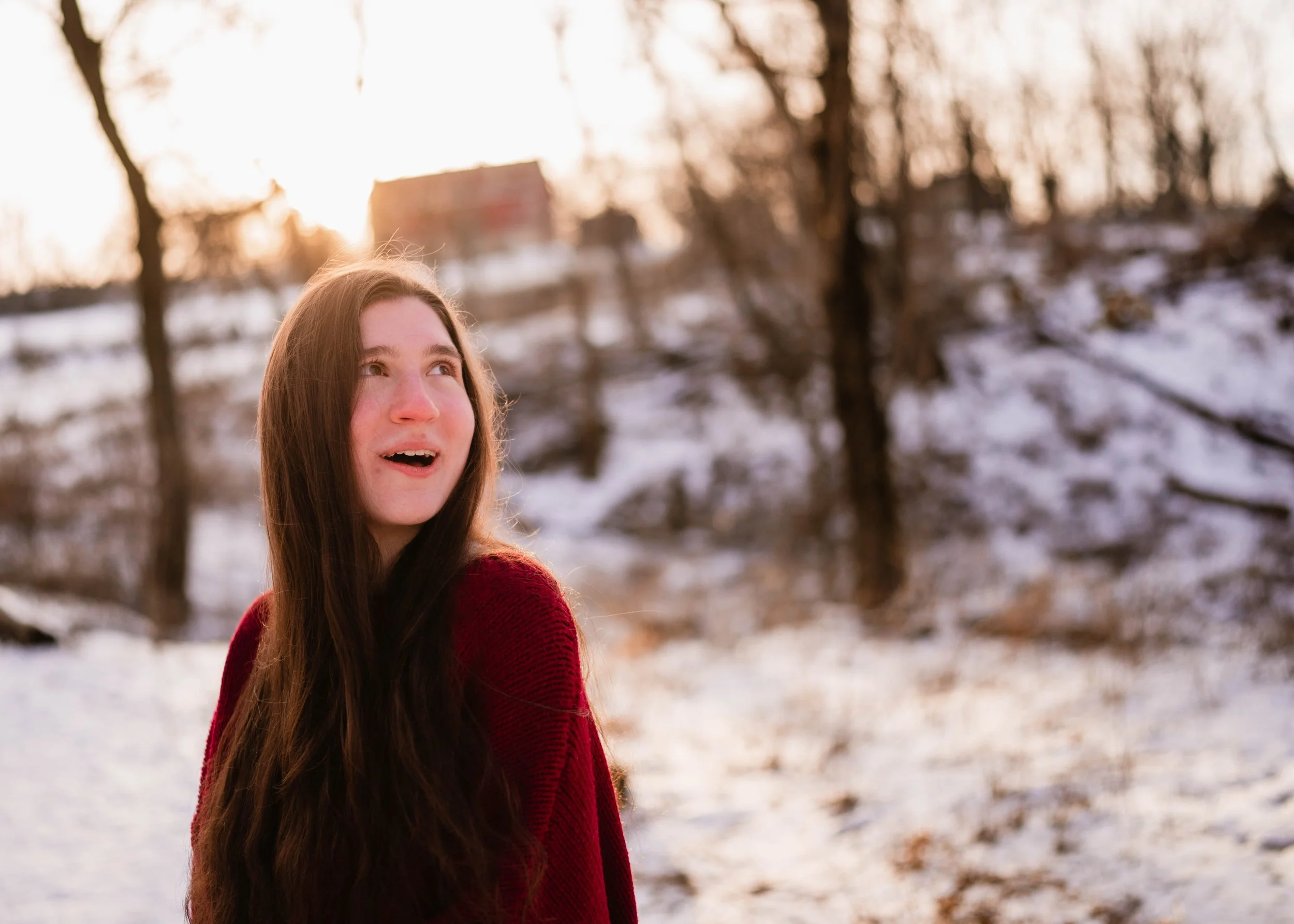 A young woman with long brown hair, wearing a red sweater, looking excited outdoors during sunset with snow on the ground and trees in the background.