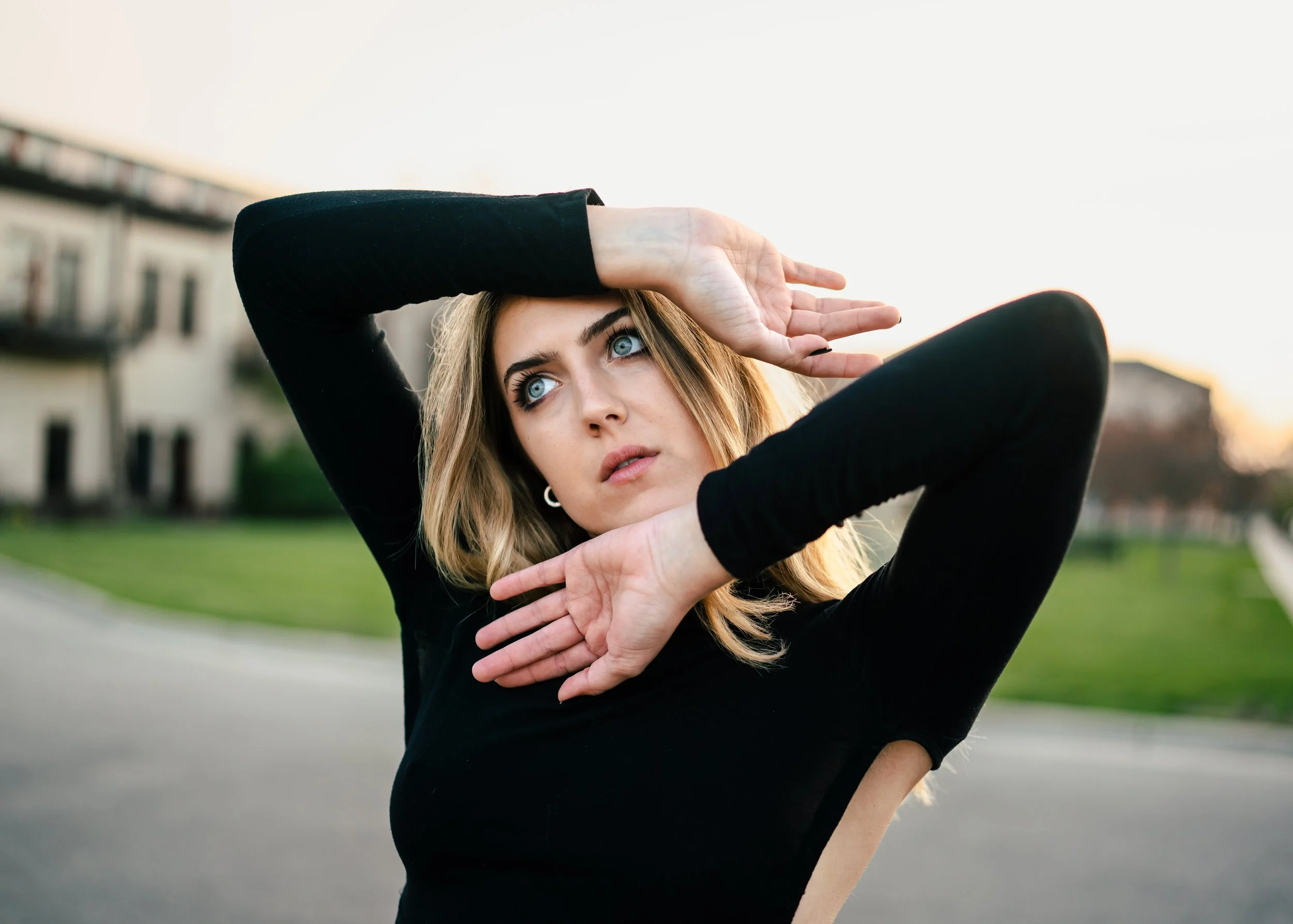 A young woman with blonde hair and blue eyes wearing a black long-sleeve shirt, outdoors during sunset, holding her hands up near her face in a dance pose, with a blurred background of buildings and greenery.