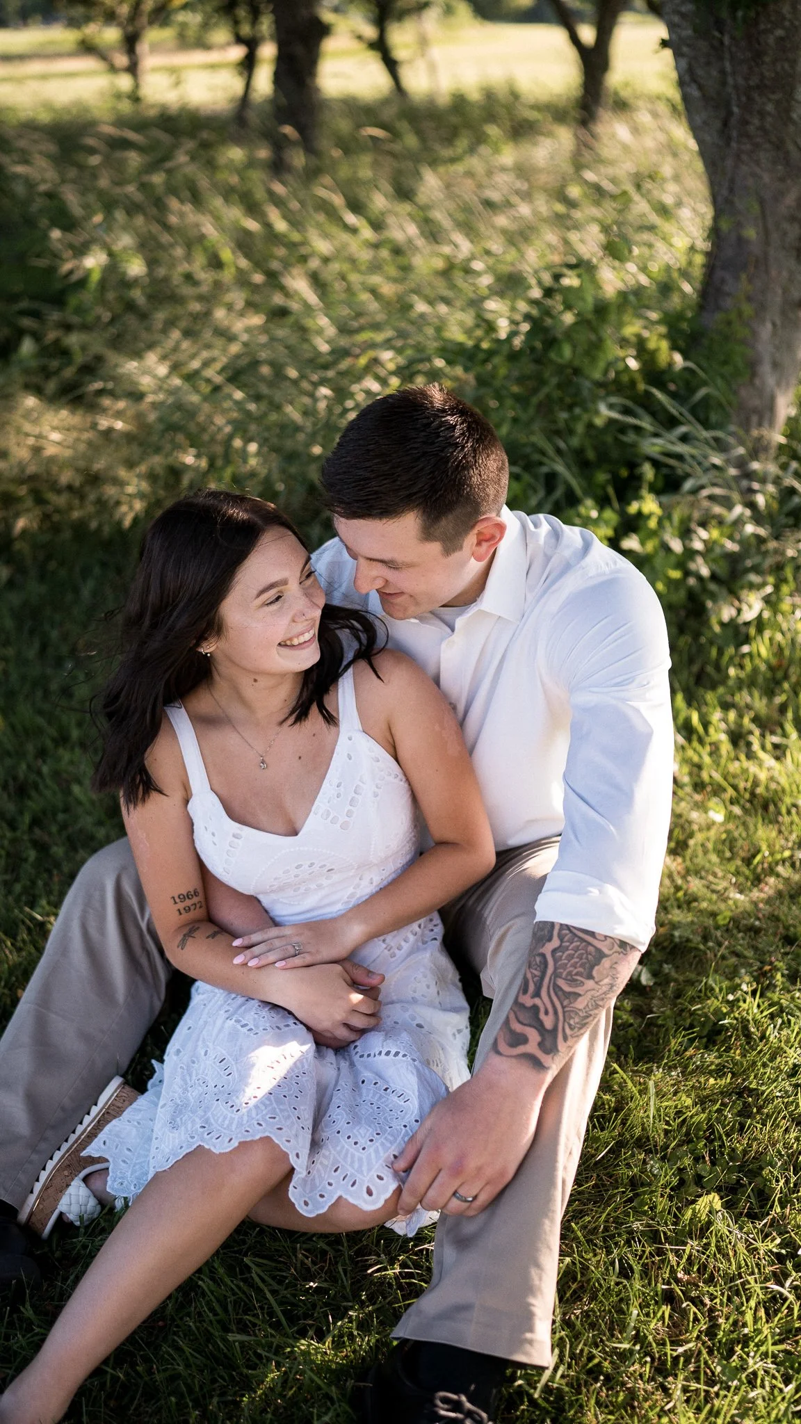 A smiling woman and a man are sitting in a grassy area outdoors, looking at each other affectionately.