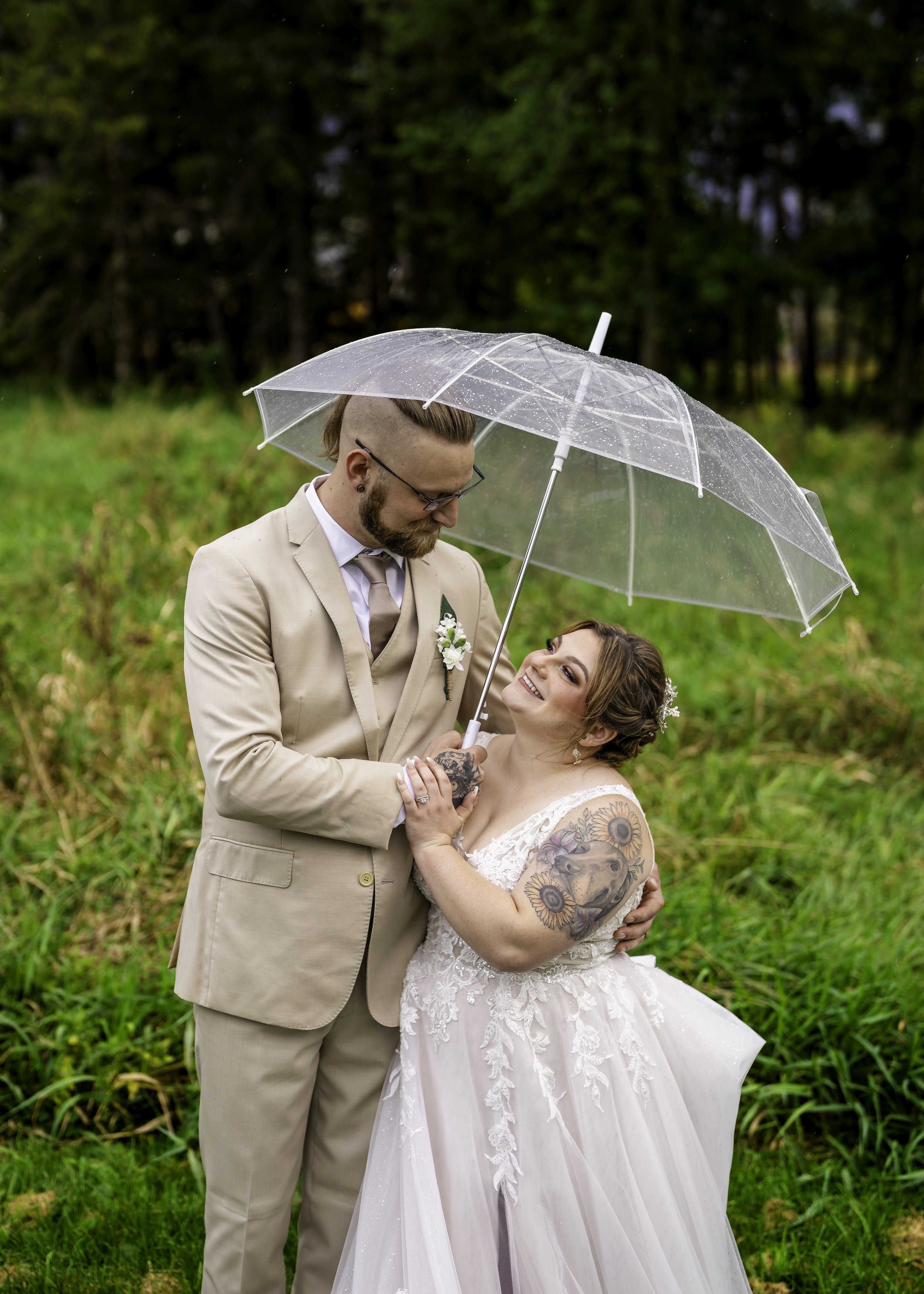 A couple on their wedding day under a clear umbrella, standing in a green outdoor setting with trees in the background. The groom wears a beige suit, while the bride wears a white lace wedding dress with colorful tattoos on her arms. They look at eac
