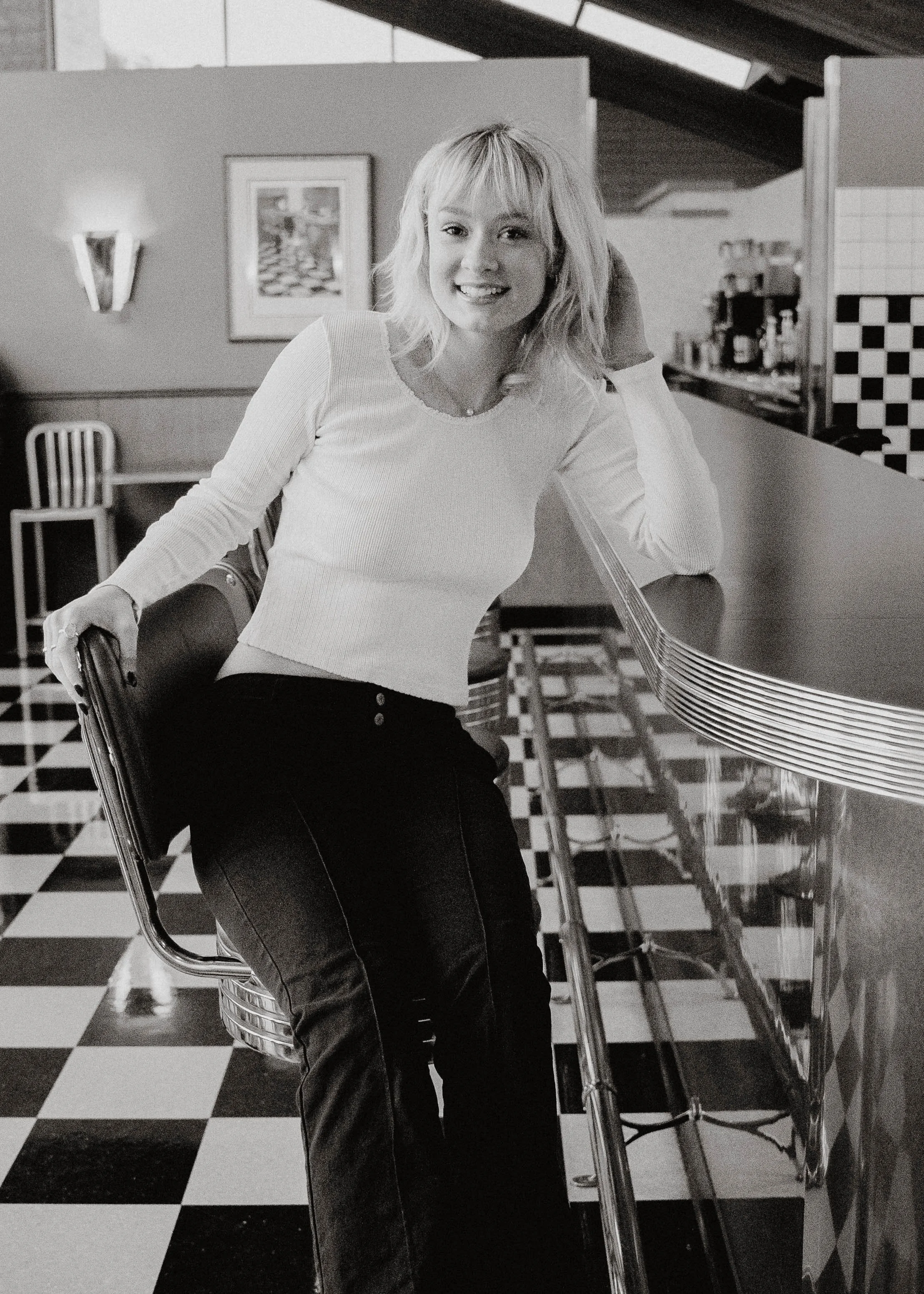 A young woman with blonde hair smiling, leaning on a counter inside a diner or cafe with checkered flooring and artwork on the wall.