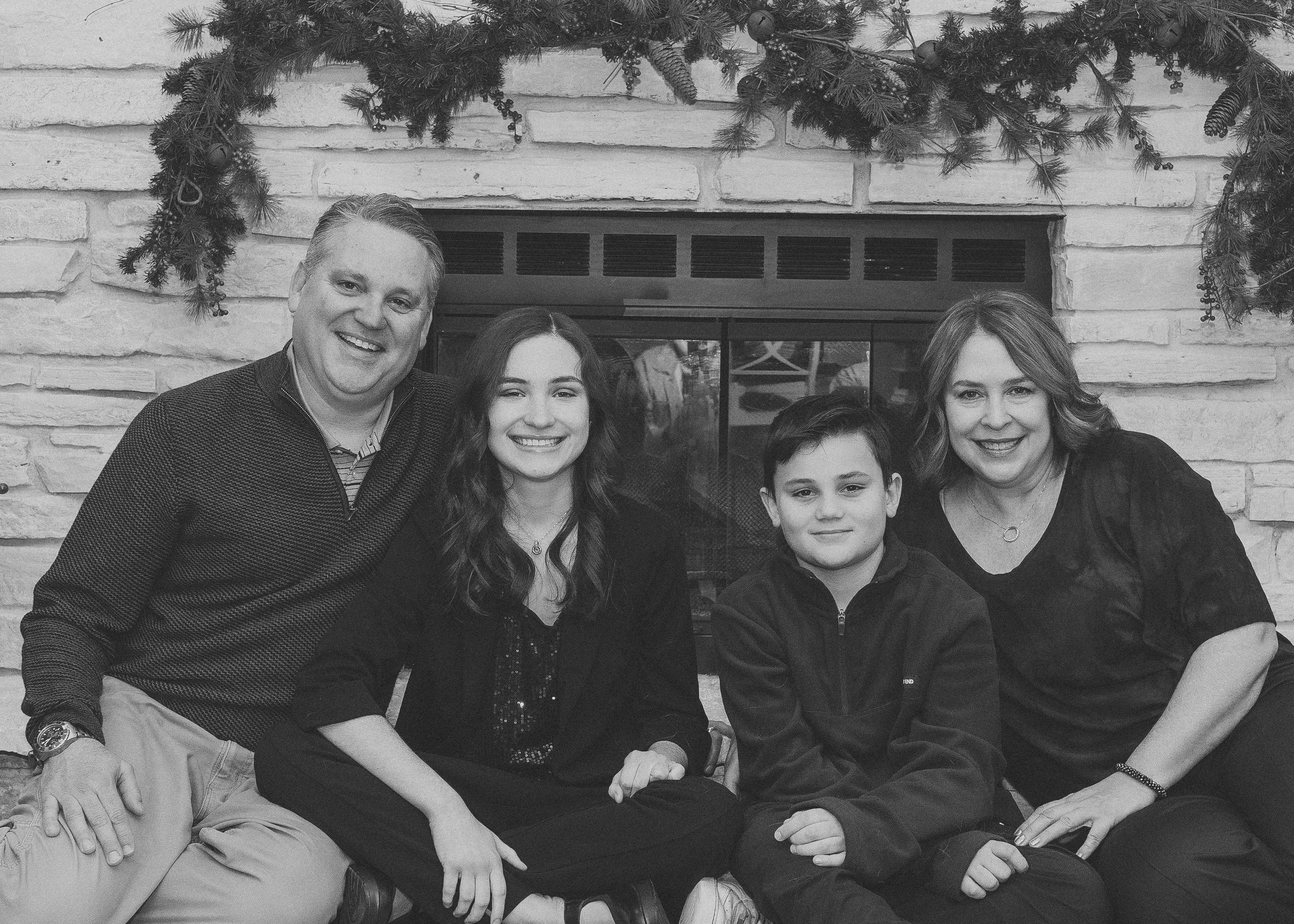A family of four sitting in front of a fireplace, smiling, with a Christmas wreath above them.