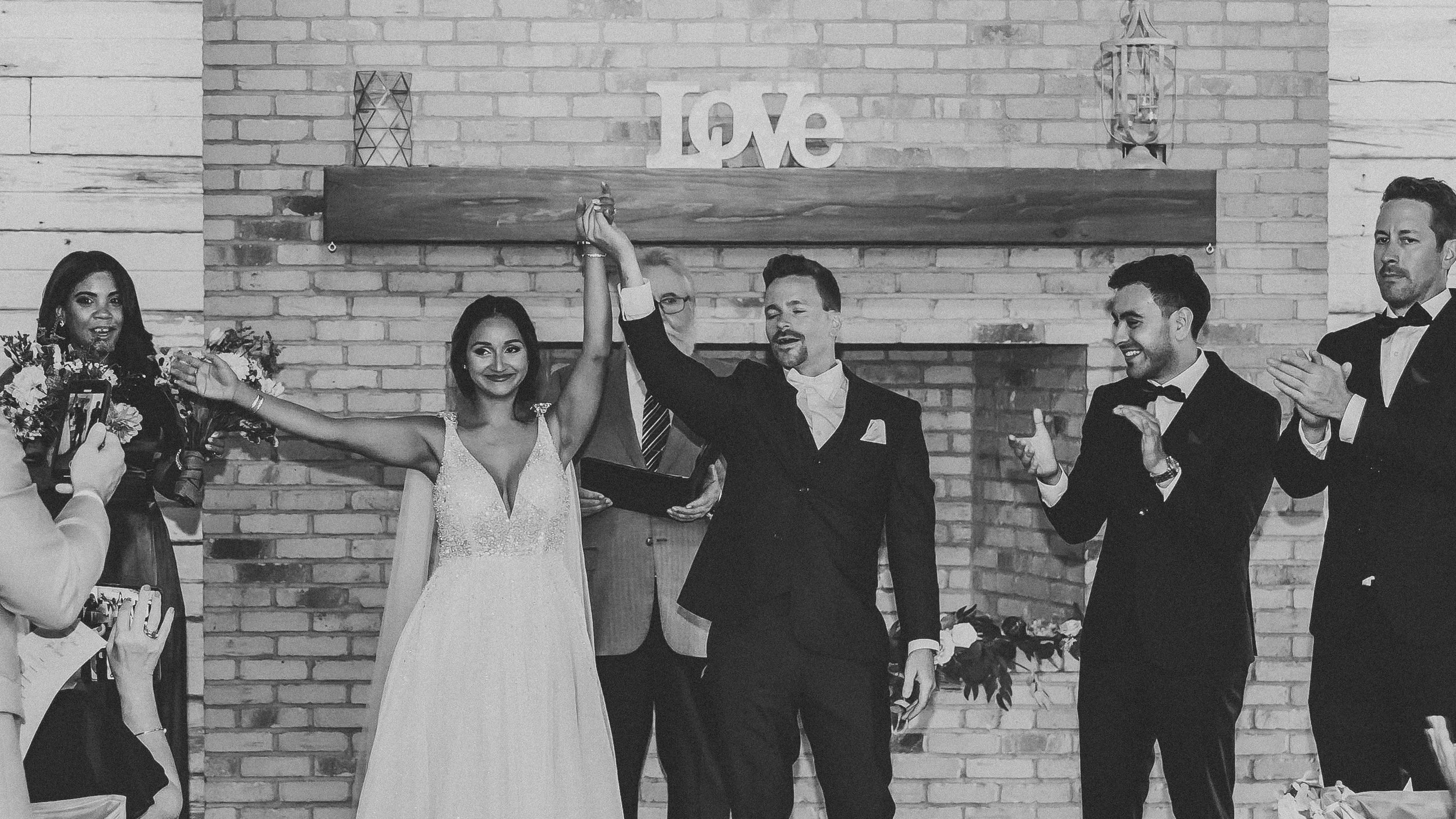 A wedding celebration with a bride and groom holding hands in front of a group of smiling guests, a brick fireplace with a decorative "LOVE" sign above, and a woman holding a bouquet of flowers to the left.