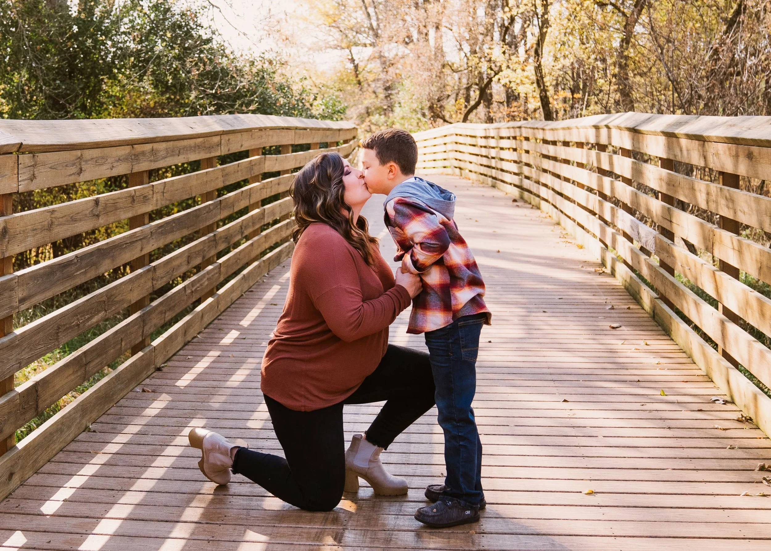 A woman kneeling on a wooden bridge, holding a young boy, as they share a kiss outdoors during autumn.