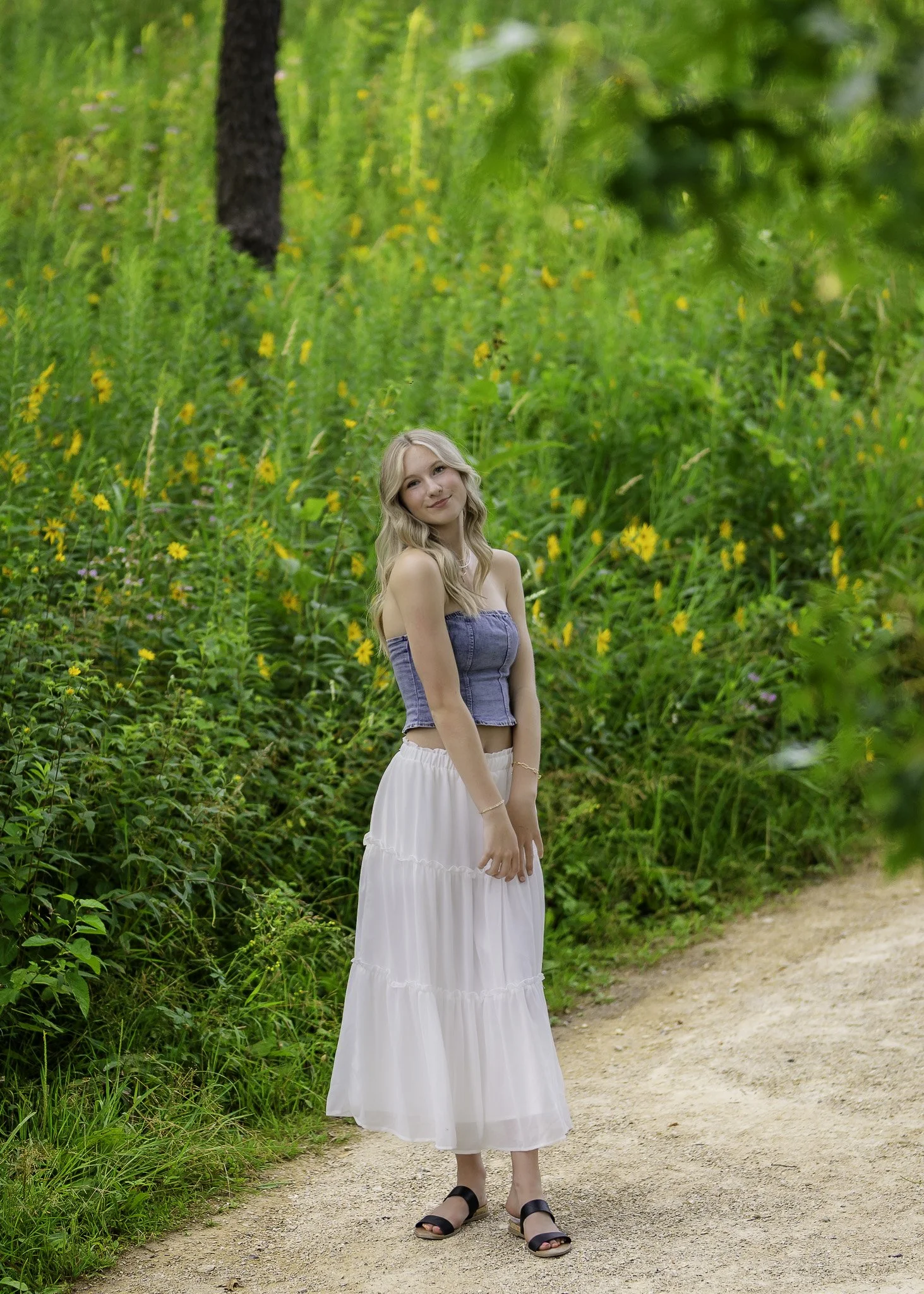 A young woman with long blonde hair standing on a dirt path in a green nature setting with yellow flowers and tall trees.