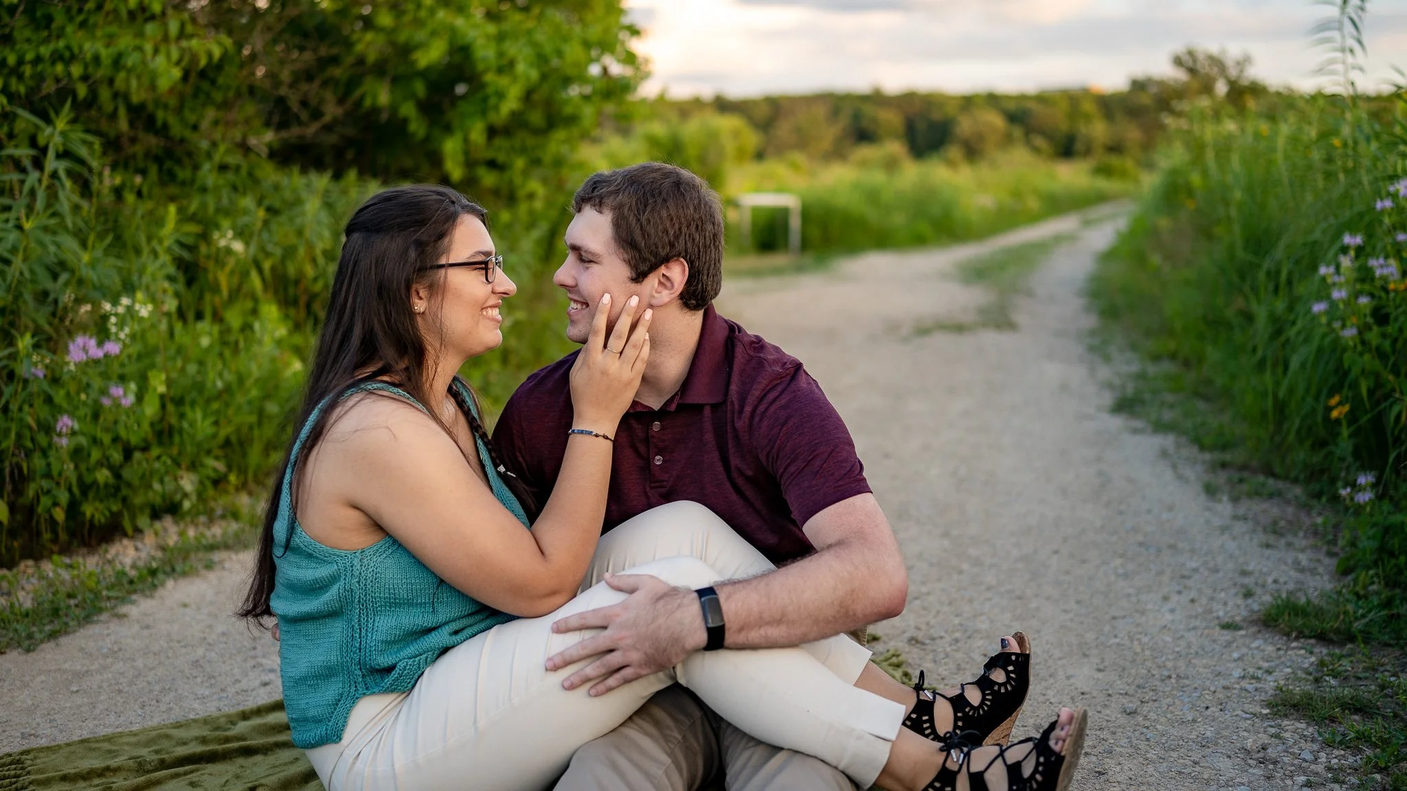 A couple sitting on a blanket on a dirt path in a green, outdoor setting during the evening, gazing at each other with smiles.