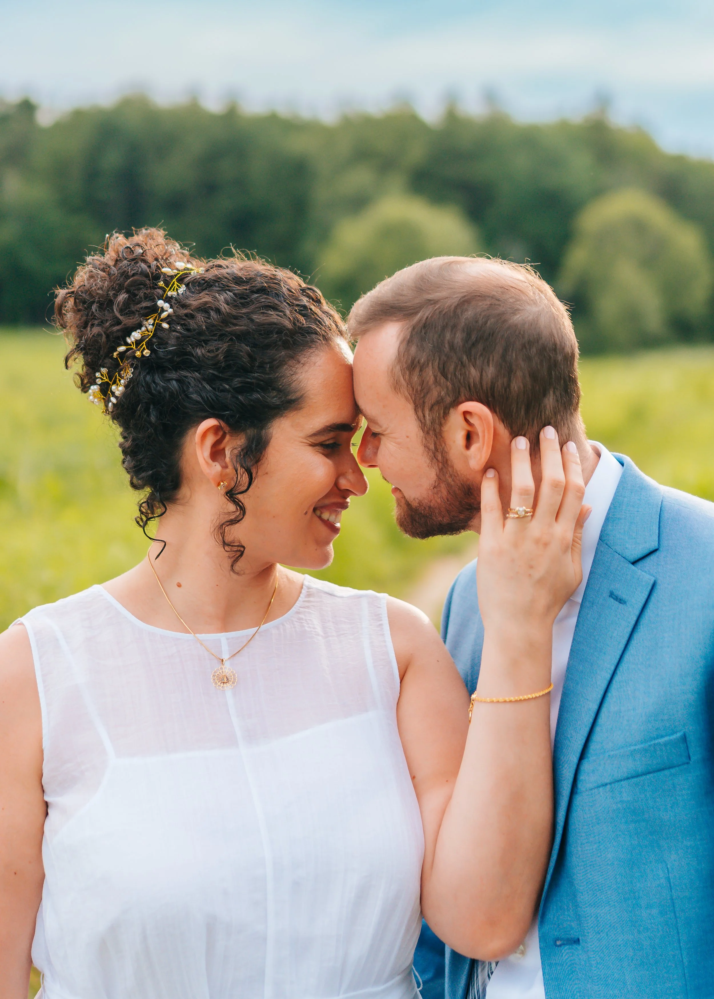 A couple in wedding attire with foreheads touching and eyes closed, smiling, outdoors with a green landscape background.