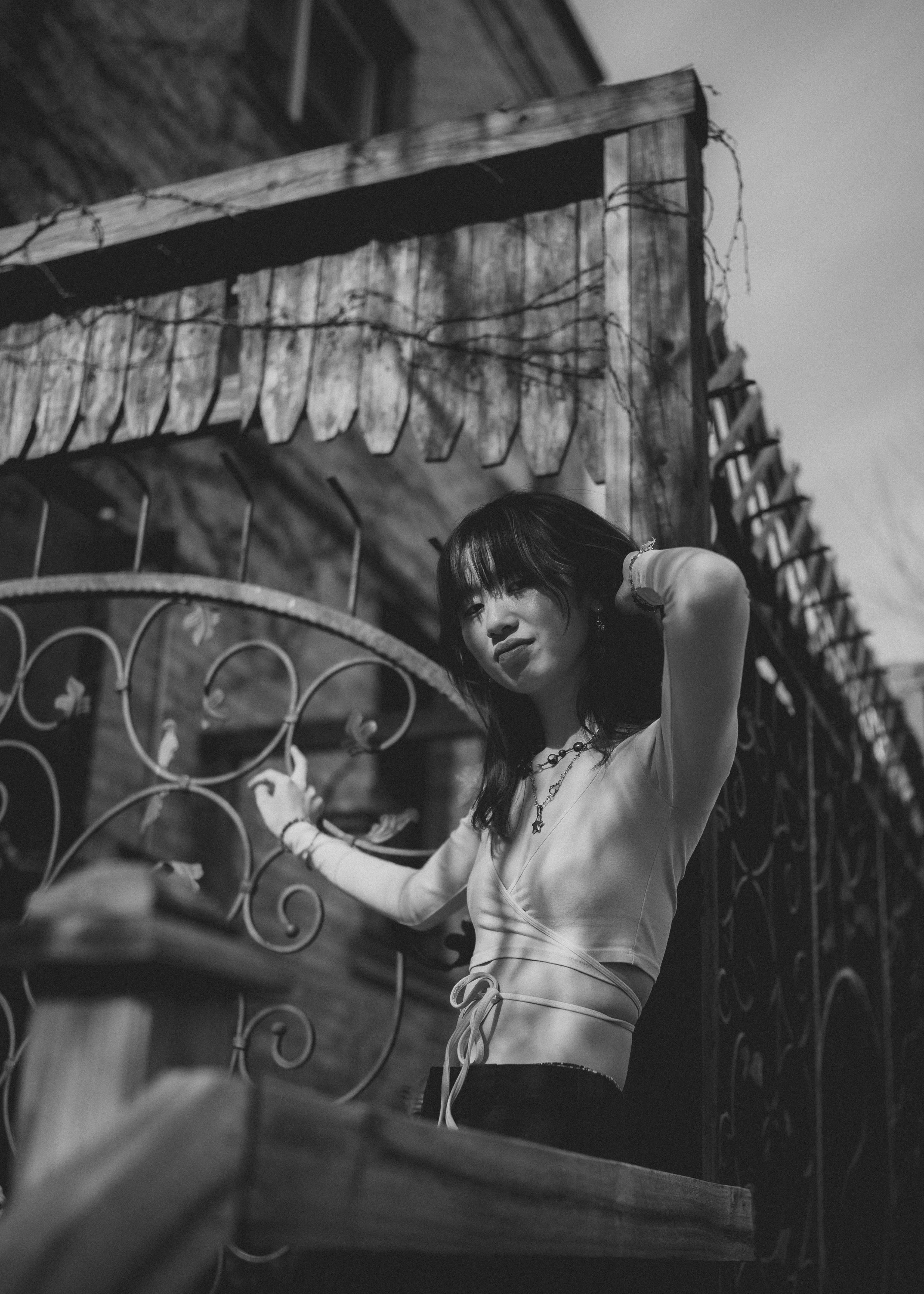 A woman with dark hair standing outdoors next to a metal fence with a decorative design, next to a wooden structure with outdoor lights, black and white photo.