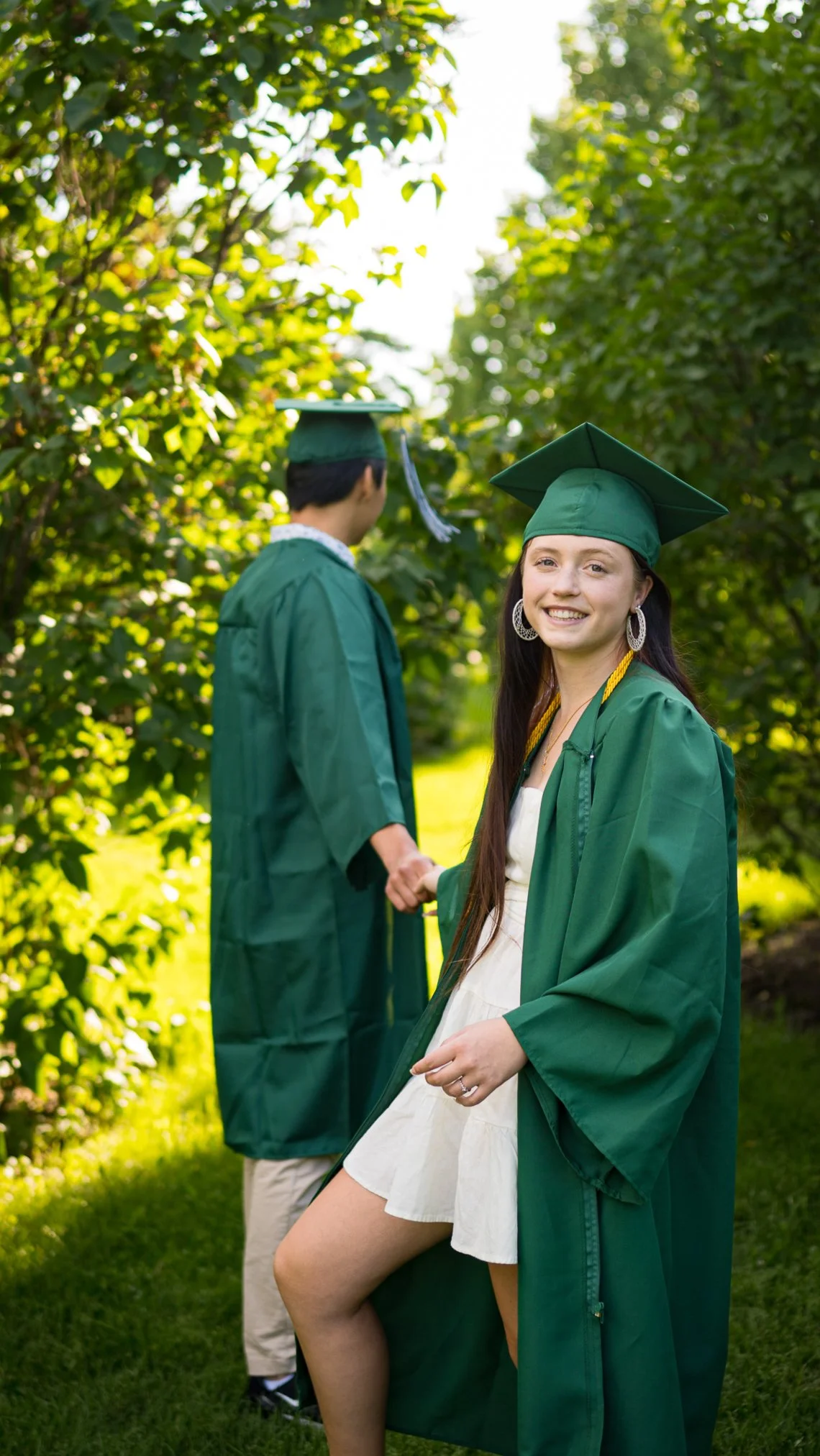 Two graduates in green caps and gowns holding hands outdoors, one smiling at the camera and wearing a white dress.