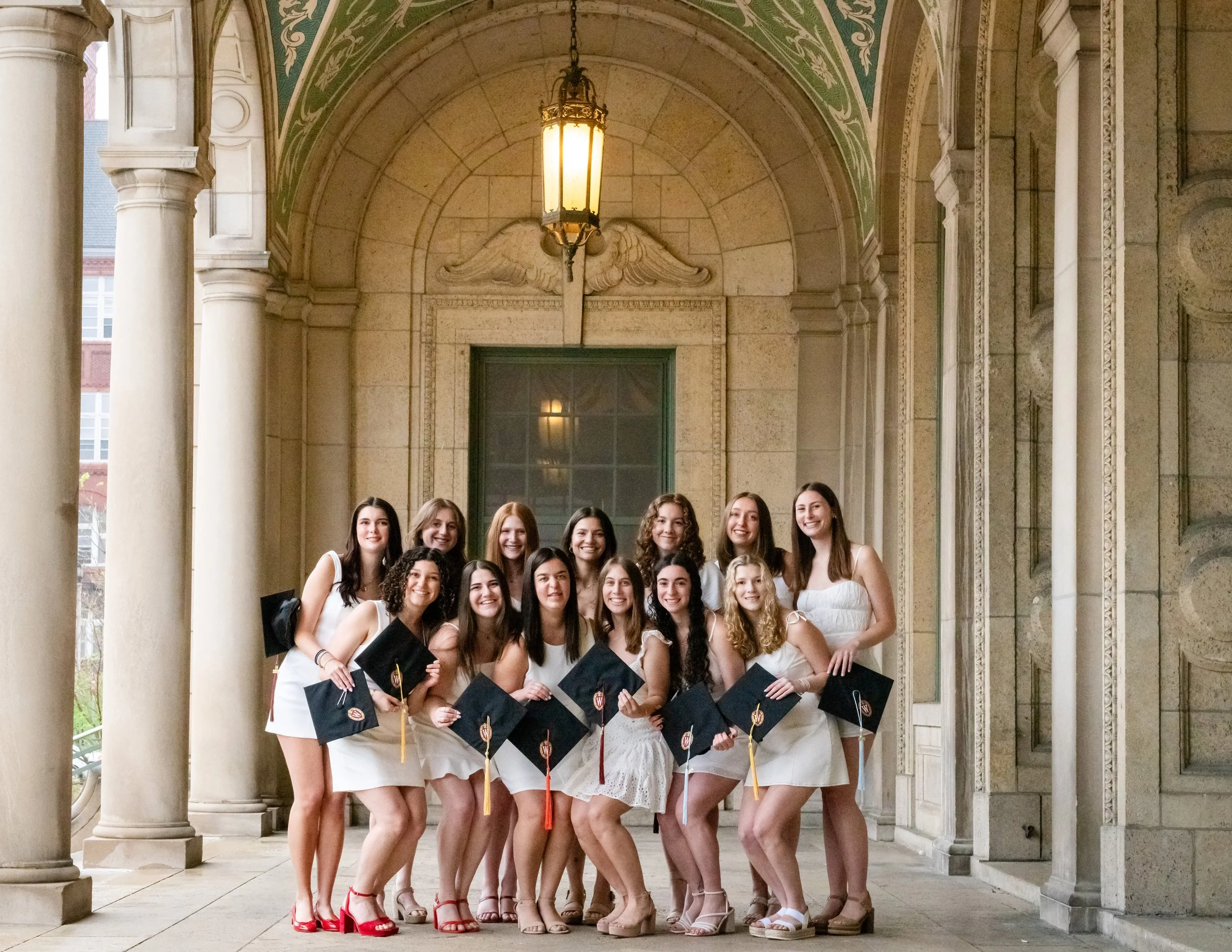 Group of young women in white dresses holding diplomas, standing under an arched, columned porch with ornate architectural details.