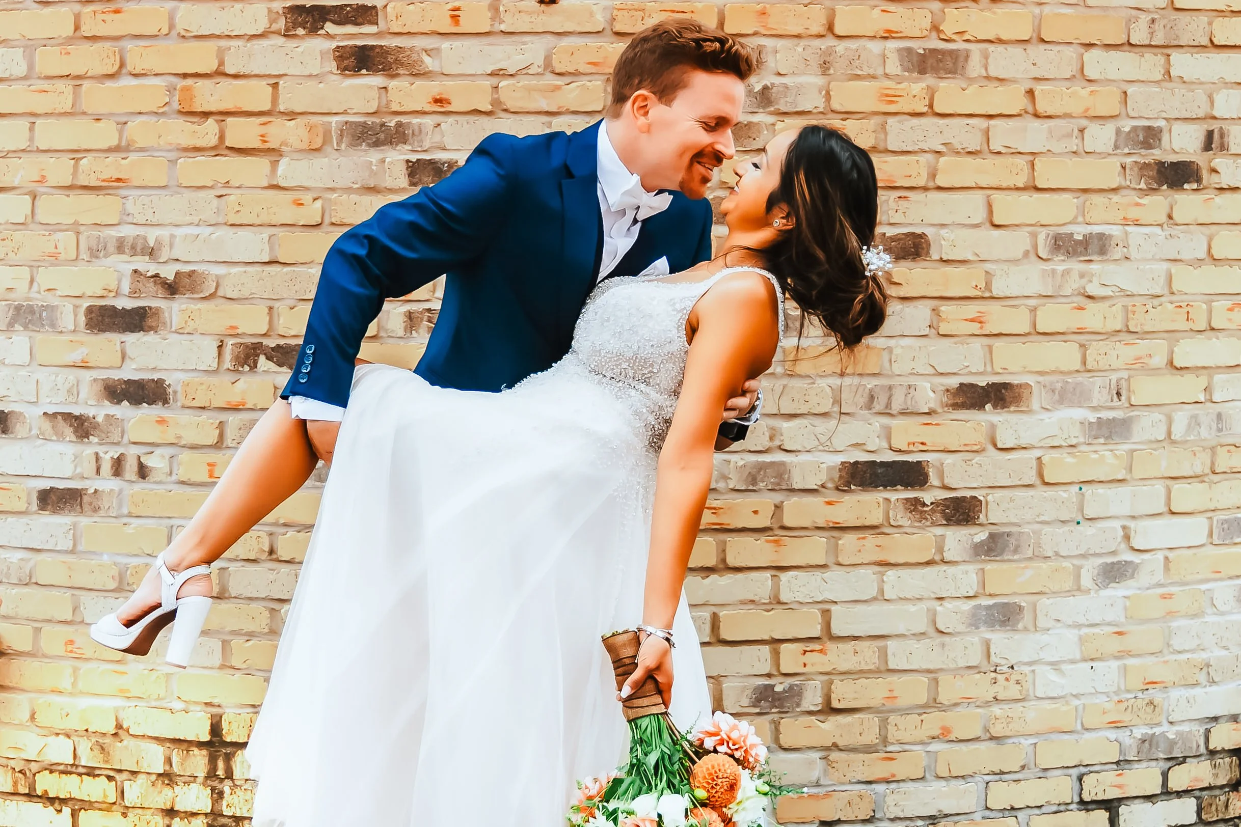 A wedding scene with a groom in a blue suit holding a bride in a white wedding dress against a brick wall, the groom leaning over the bride, who holds a bouquet of flowers.