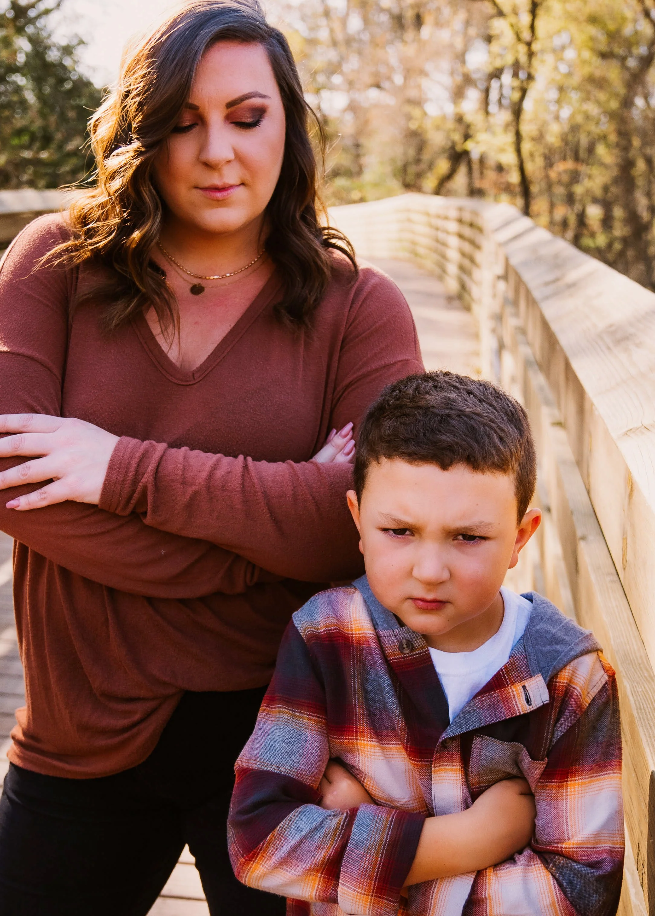 A woman with brown hair and a young boy with crossed arms standing outdoors on a wooden bridge, with trees and fall foliage in the background.