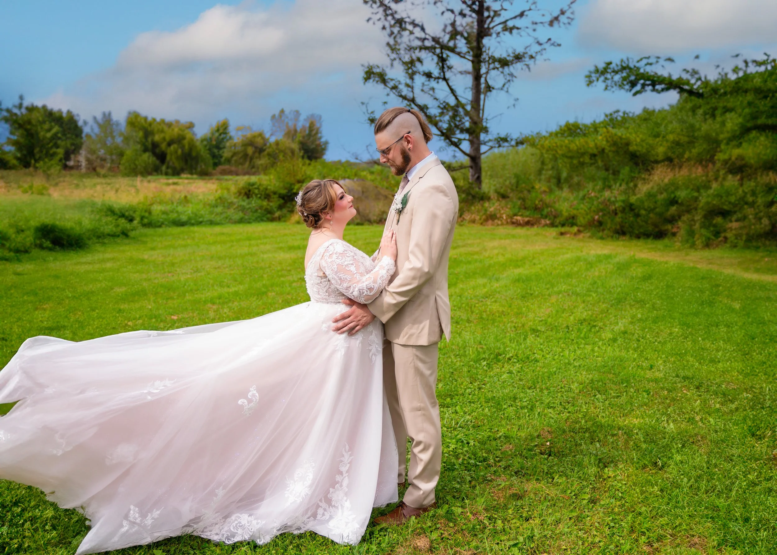 Bride and groom standing on green grass in a field, holding hands and looking at each other, with trees and a partly cloudy sky in the background.