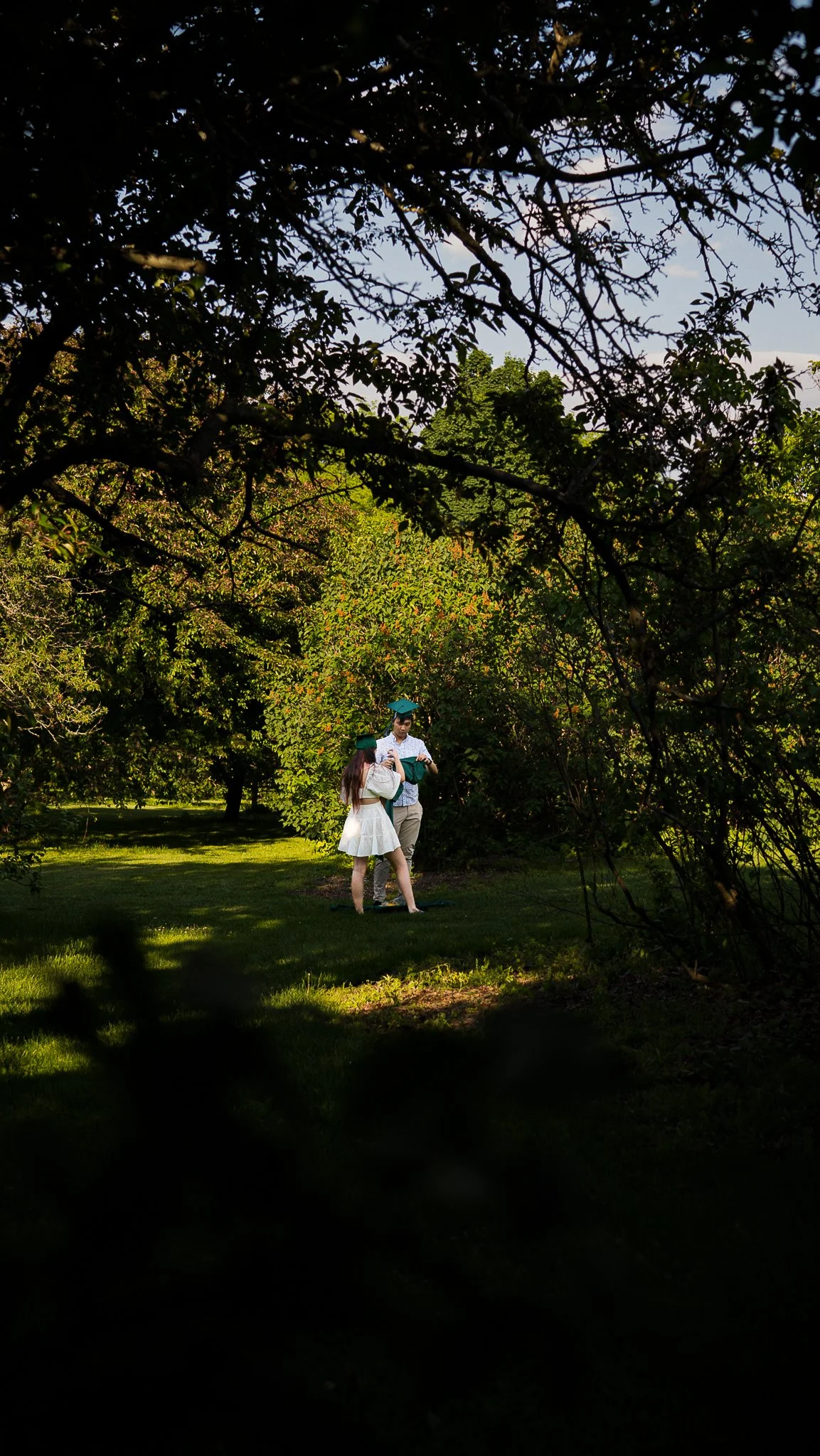 Two graduates in caps and gowns standing together outdoors in a park, surrounded by trees and greenery, with the sky visible in the background.