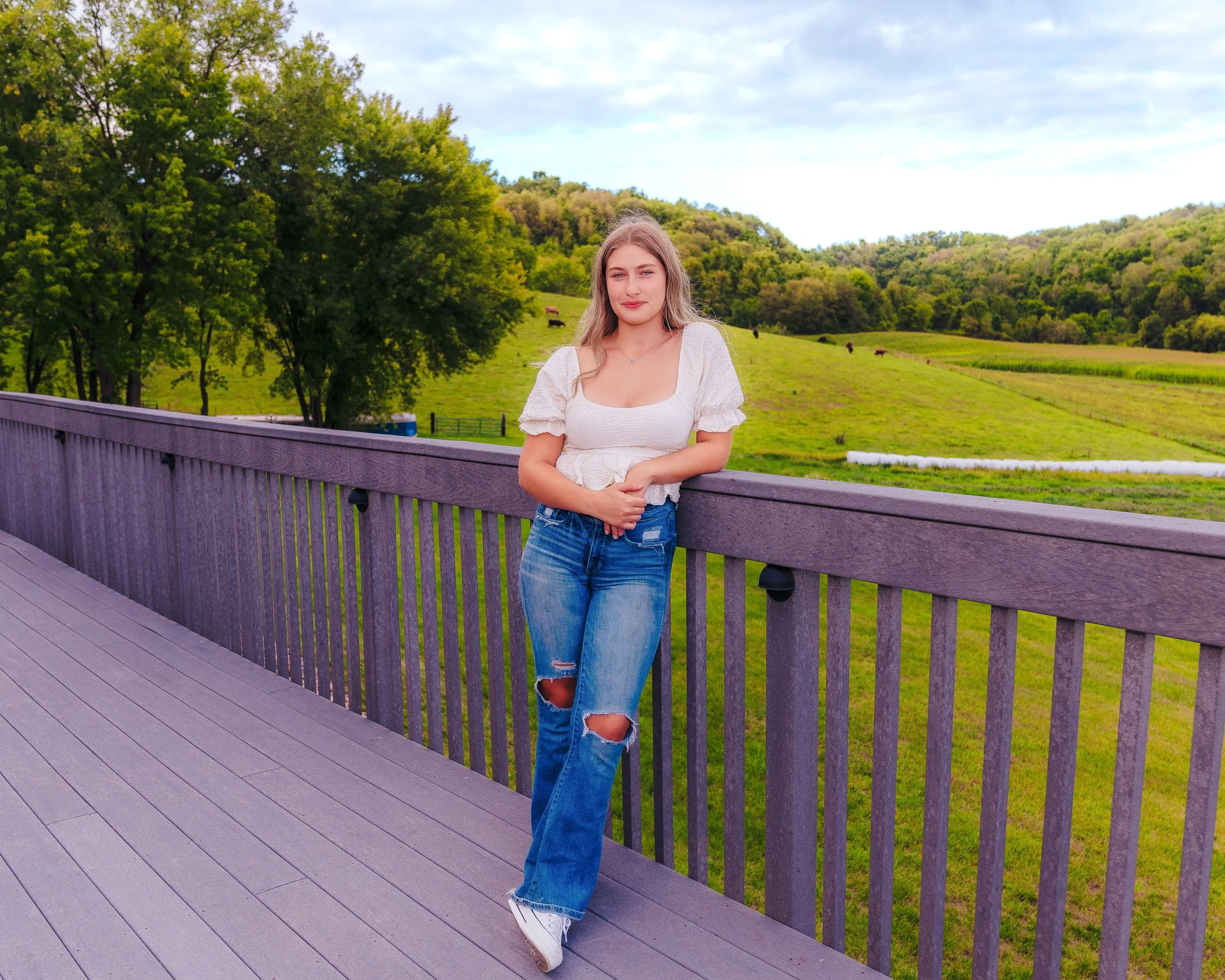 A young woman with blonde hair wearing a white top and ripped blue jeans leaning against a wooden railing on a scenic outdoor deck, with green fields, trees, and rolling hills in the background.