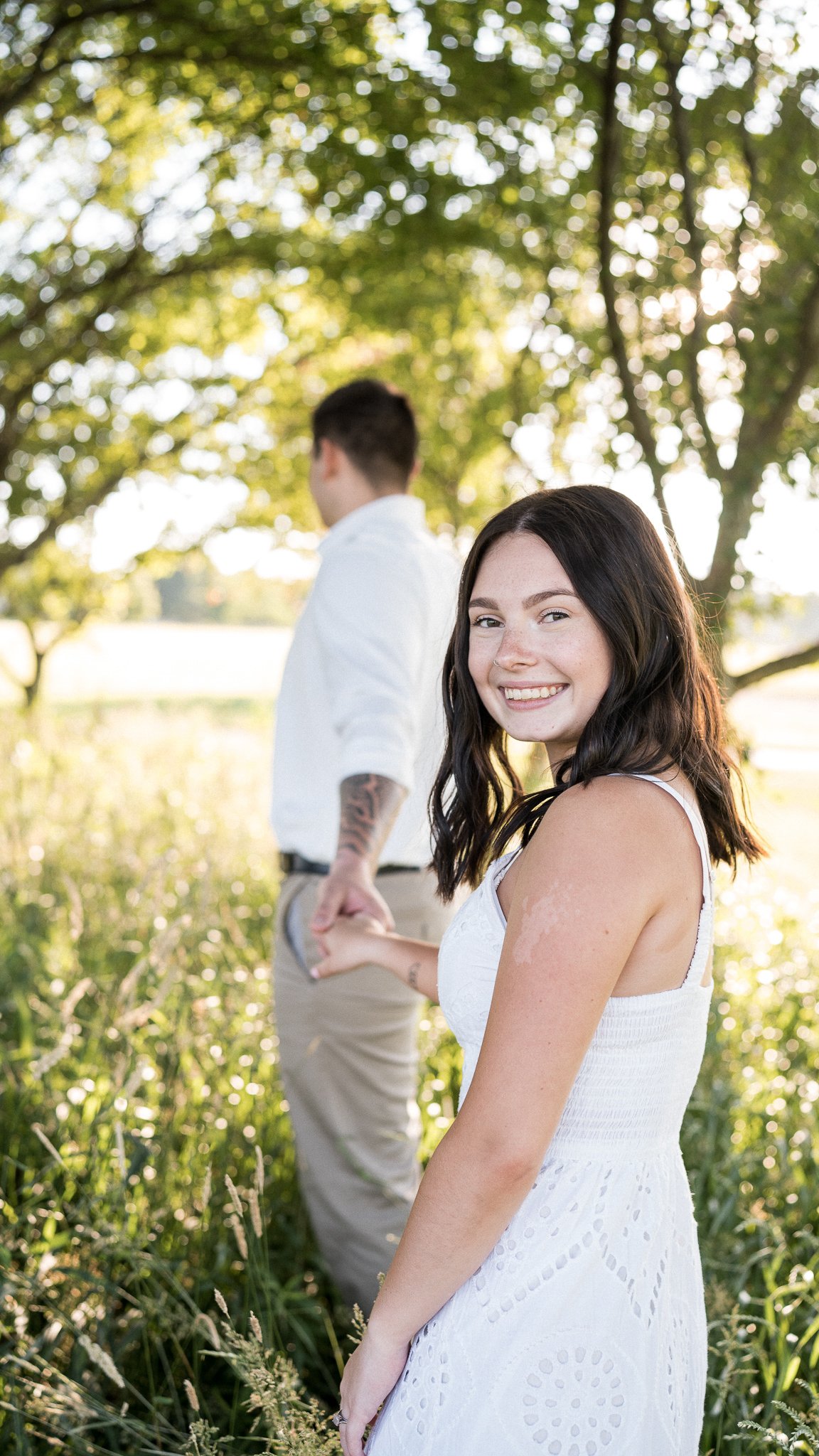 A young woman in a white dress smiling at the camera while holding hands with a young man in a white shirt in a lush outdoor setting with green trees and sunlight filtering through the leaves.