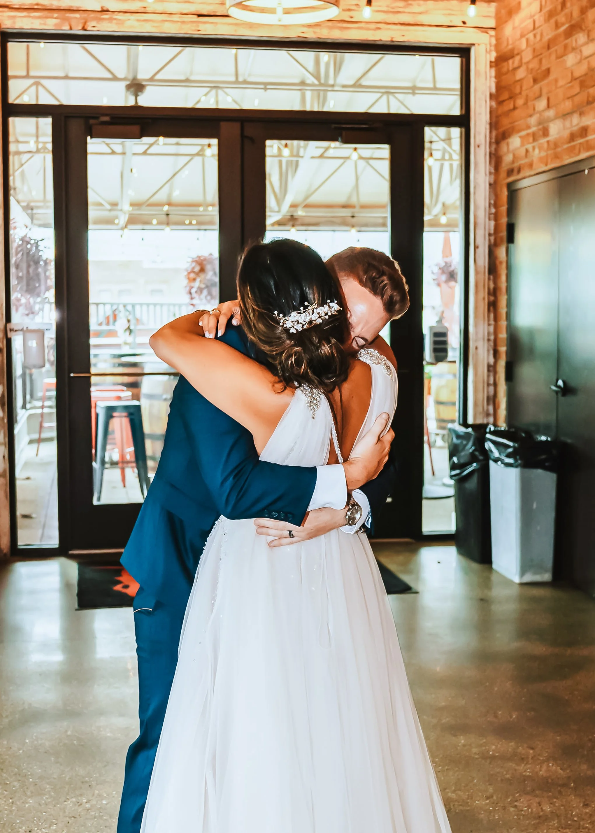 A bride and groom embrace inside a venue, with the bride in a white wedding dress and the groom in a navy blue suit. The bride has dark hair with an accessory, and the groom has light brown hair.