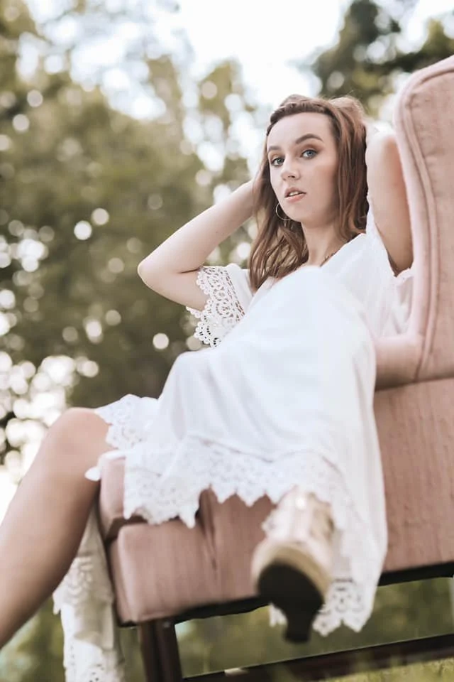 A young woman with brown hair and hoop earrings reclining on a pink vintage armchair outdoors, wearing a white lace dress, with trees in the background.